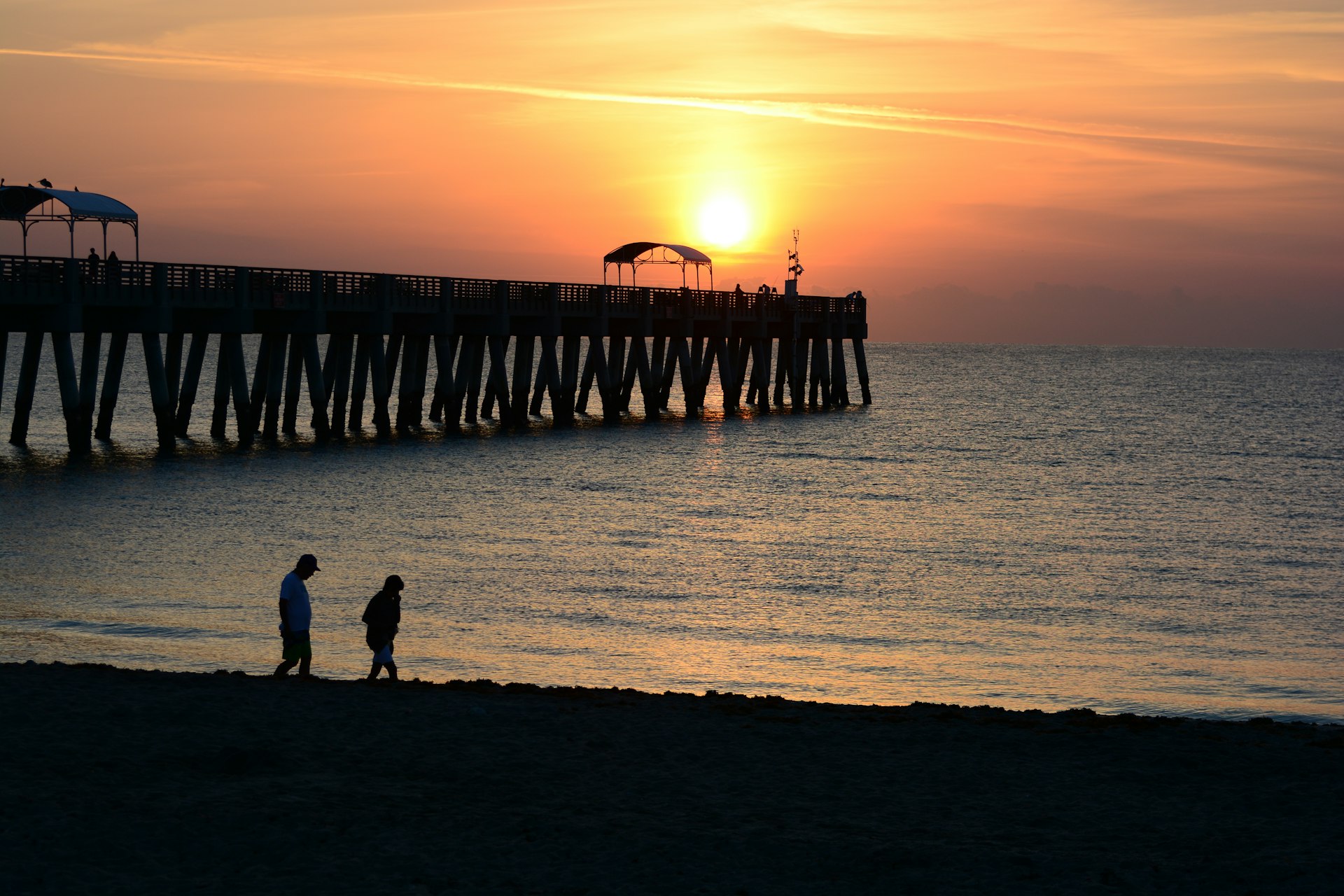 A sunrise over a beach pier with the silhouettes of two adults walking on the beach