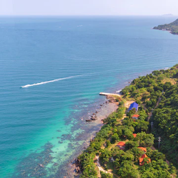 View over Hon Thom Island coast from Phu Quoc cable car with a speed boat down on the sea and rainforest with som small hauses and the horizon in background.