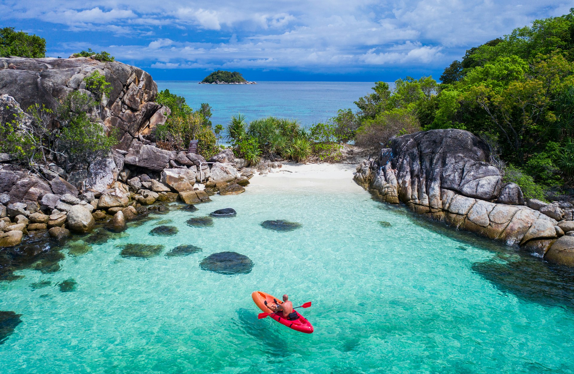 A man sits in a small red kayak floating in a turquoise bay sheltered by large rocks