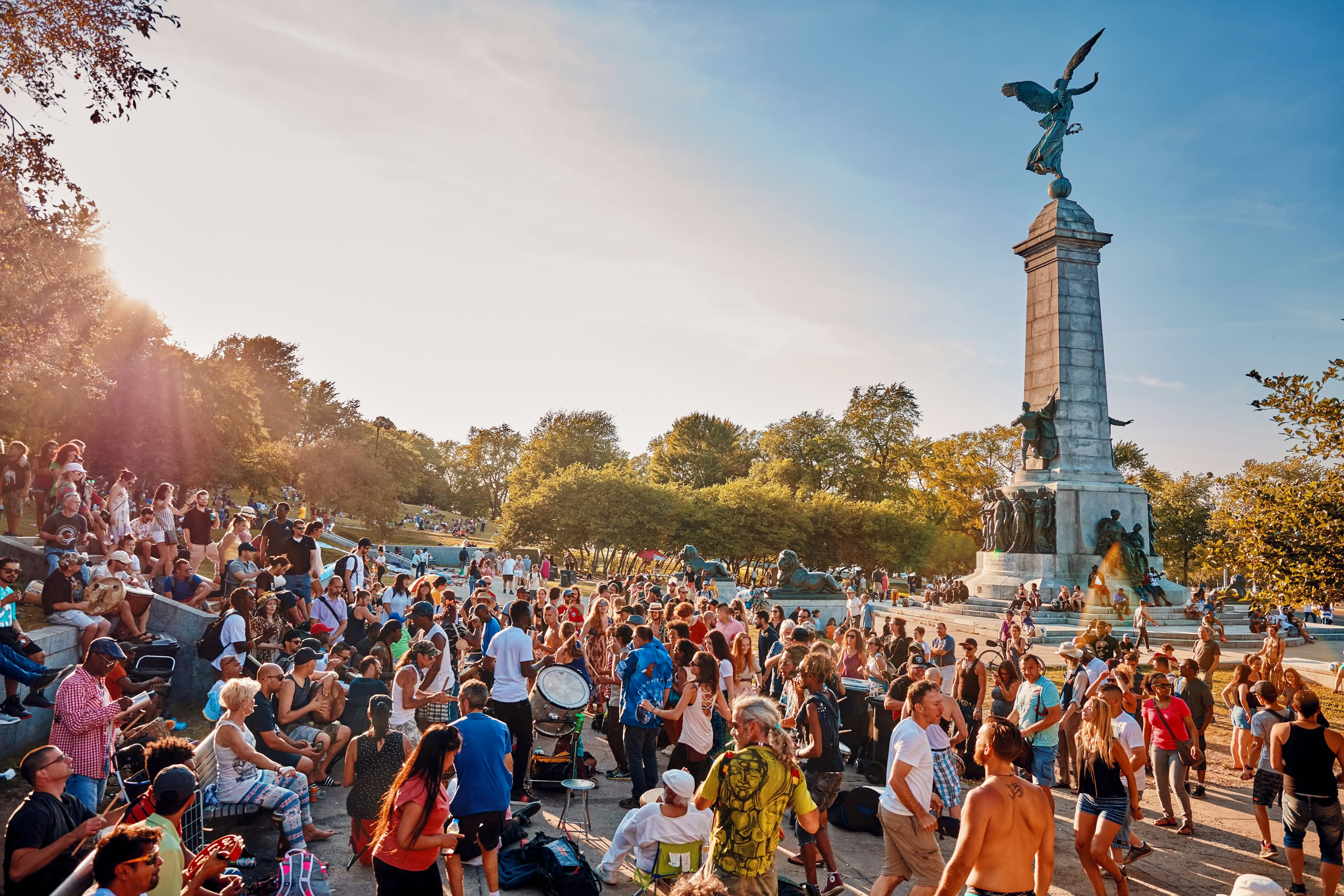 A diverse crowd of people gathered and cheering around a tall monument in a park. Some people are beating drums