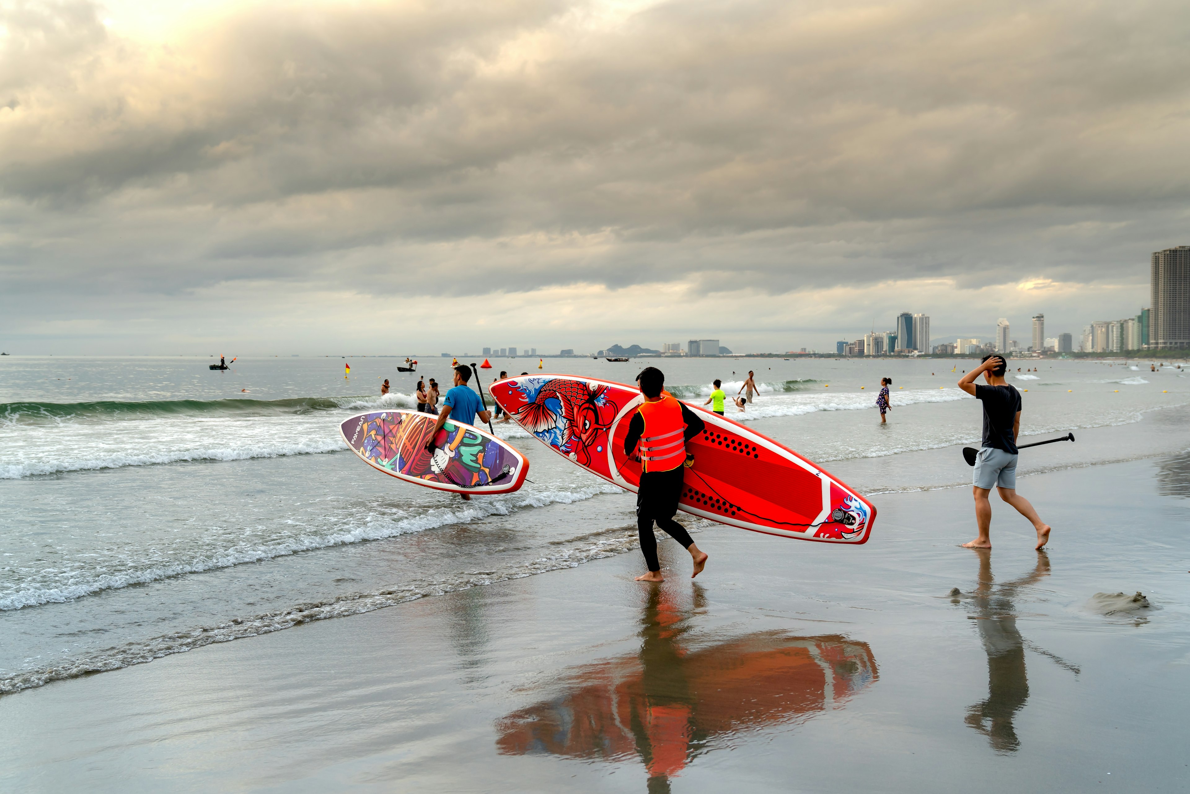 People holding surfboards walk into the gently breaking waves at a city beach with the towers of Danang city visible in the distance