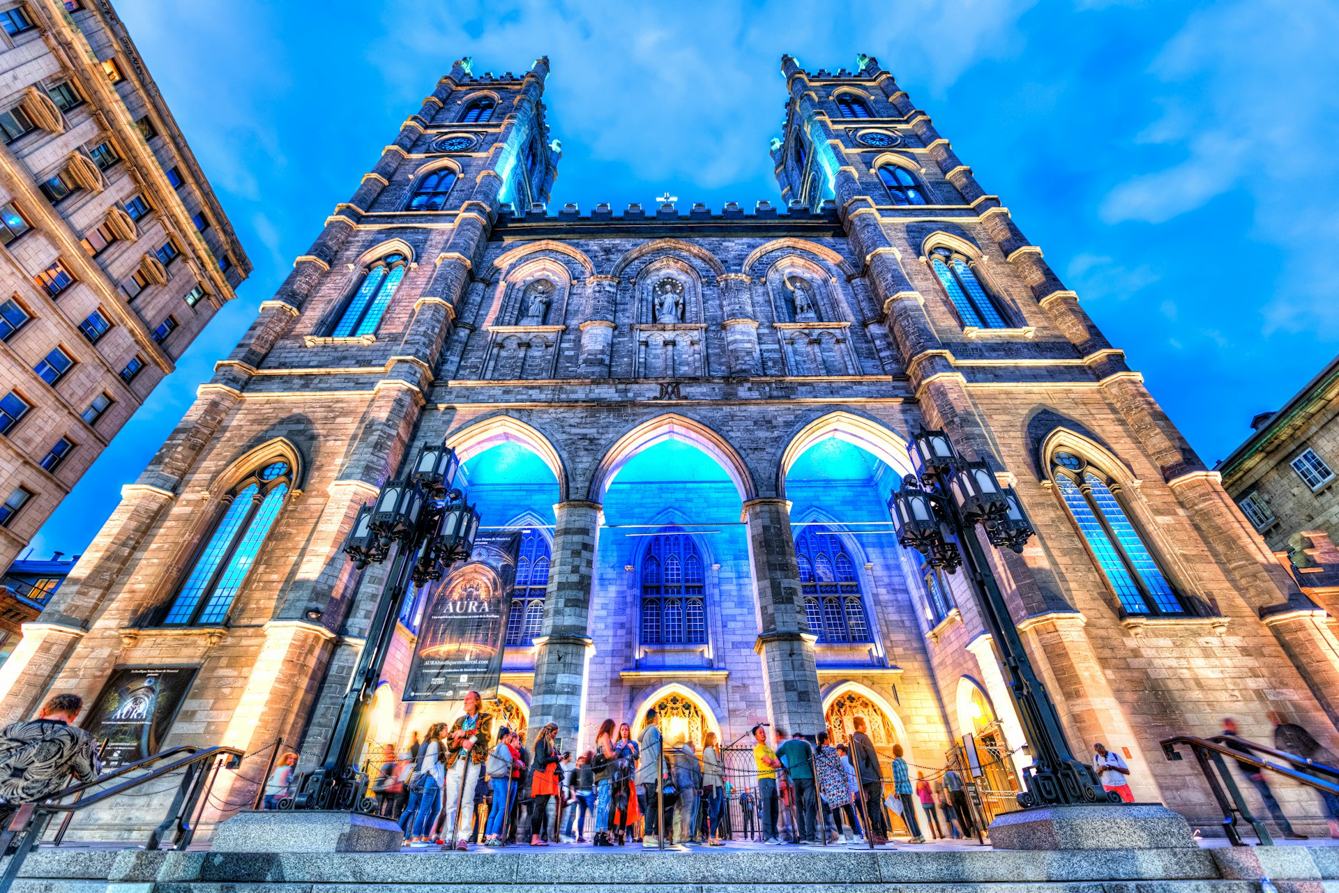 People gather outside a Gothic cathedral in the evening, which is lit up with pale blue lights