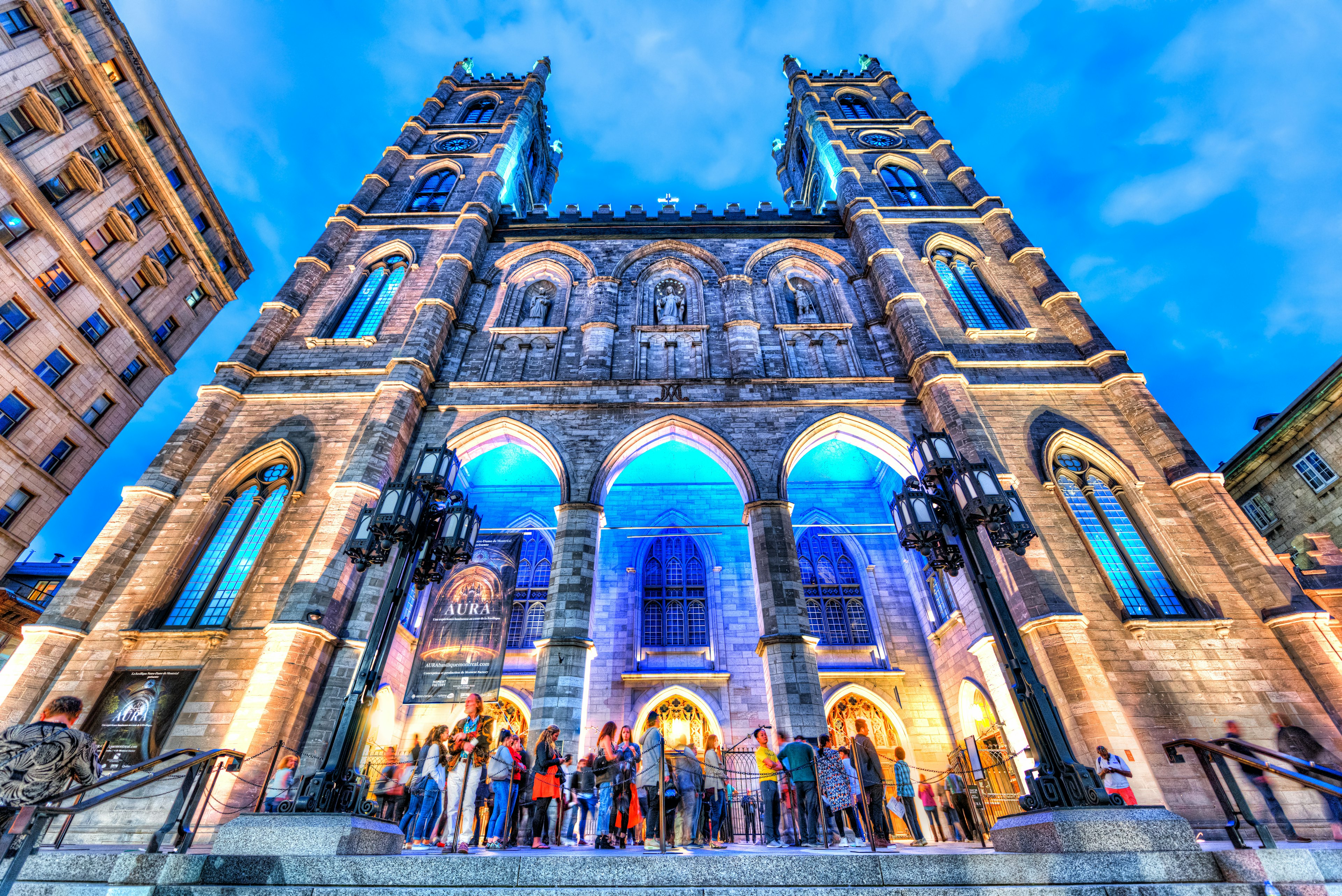 People gather outside a Gothic cathedral in the evening, which is lit up with pale blue lights