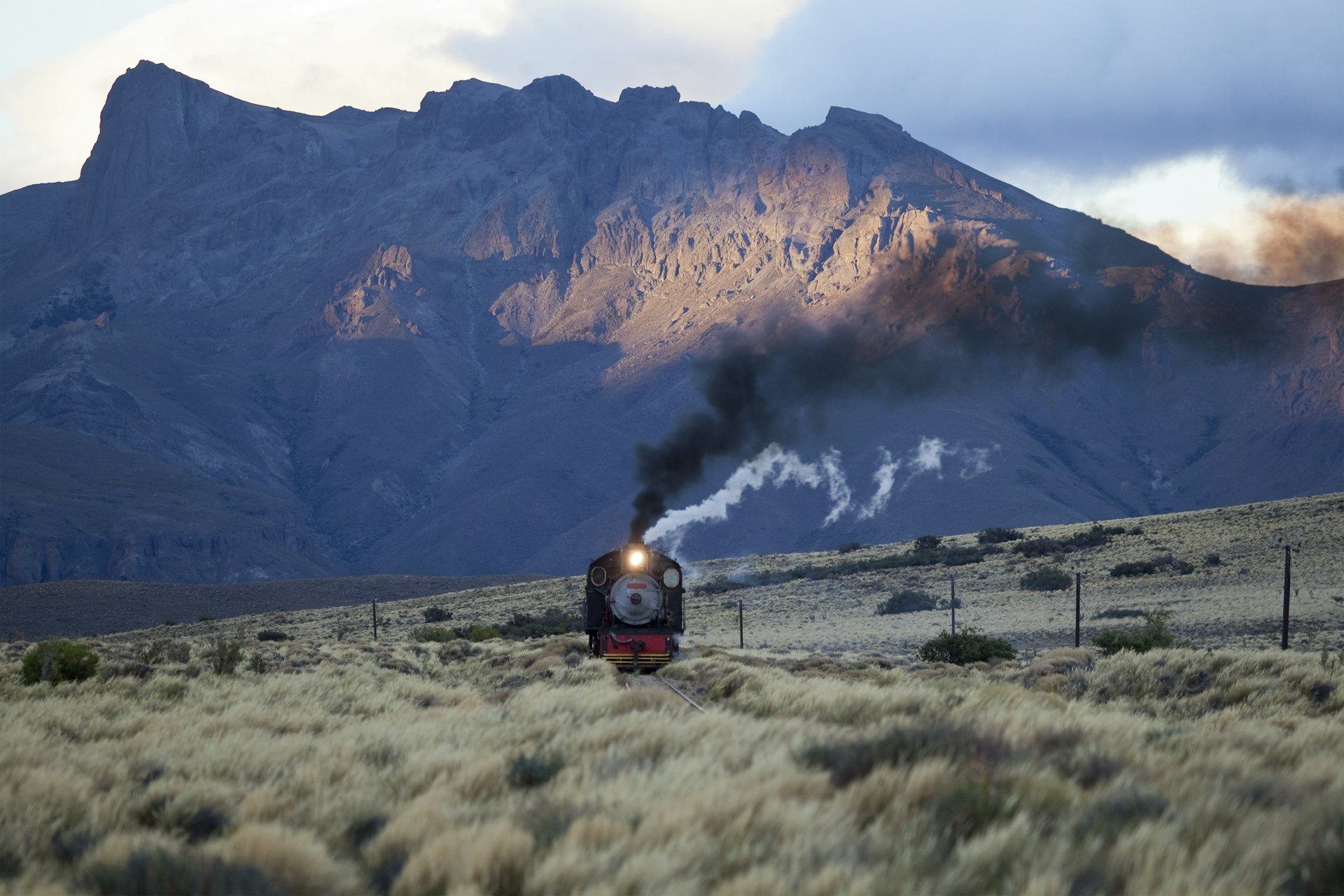 An old steam train traveling through the Patagonian landscape with grass in the foreground and mountain peaks behind.