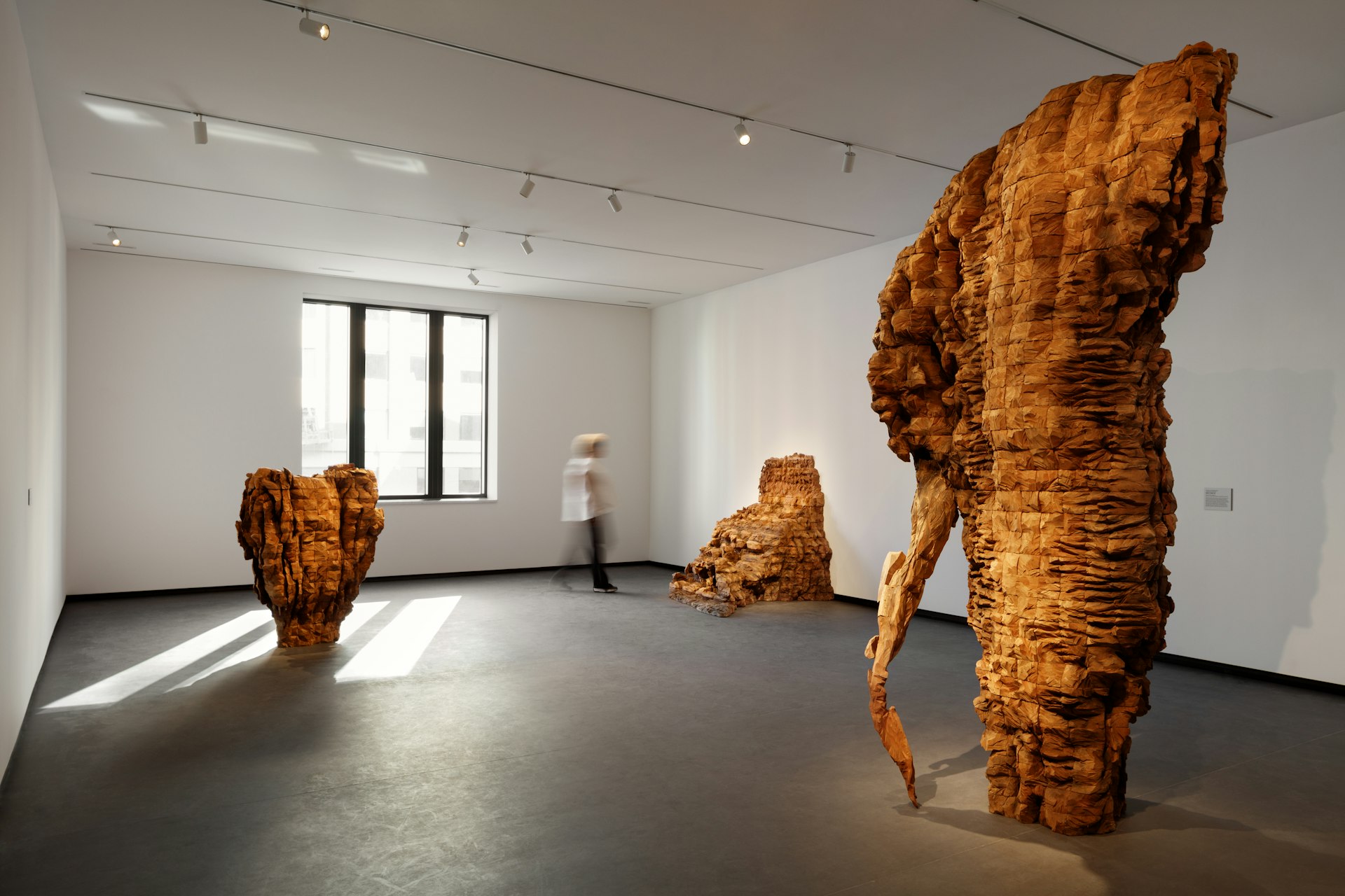 A visitor walks in the middle of a modern museum gallery. Three abstract sculptures made from beams of cedar are stacked and roughly carved.