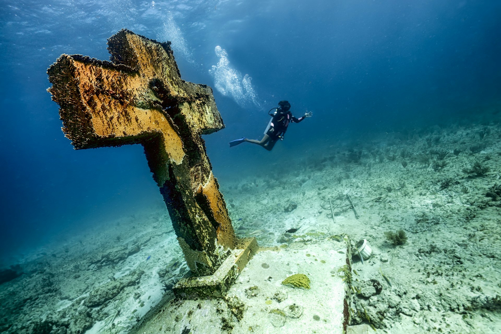 A scuba diver swims past a cross statue on the seabed at Cruz de la Bahia on bottom of sea, Isla Mujeres, Caribbean Sea.