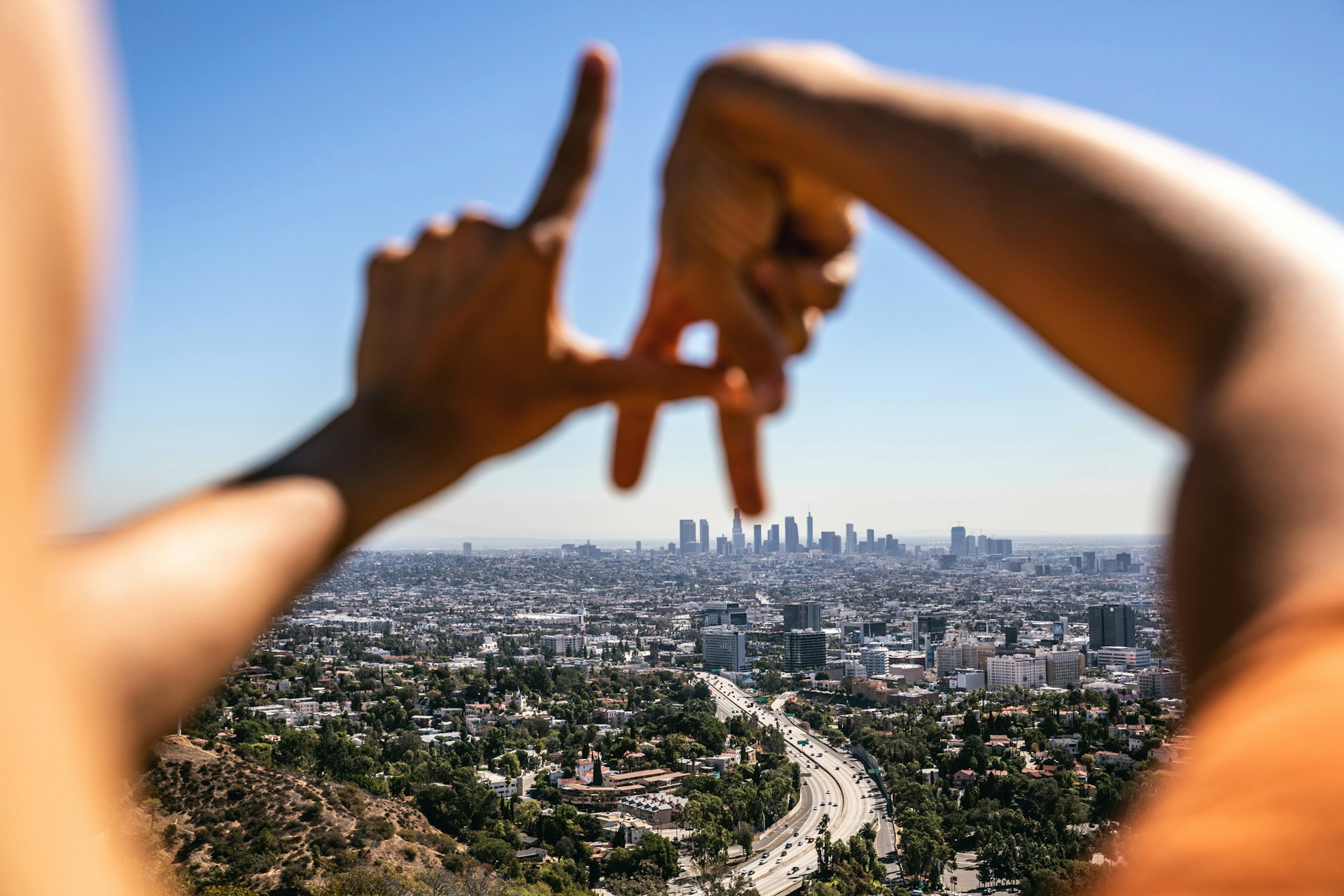 A Hispanic woman holds her hands in the shape of 'LA' whilst looking across the cityscape of Los Angeles