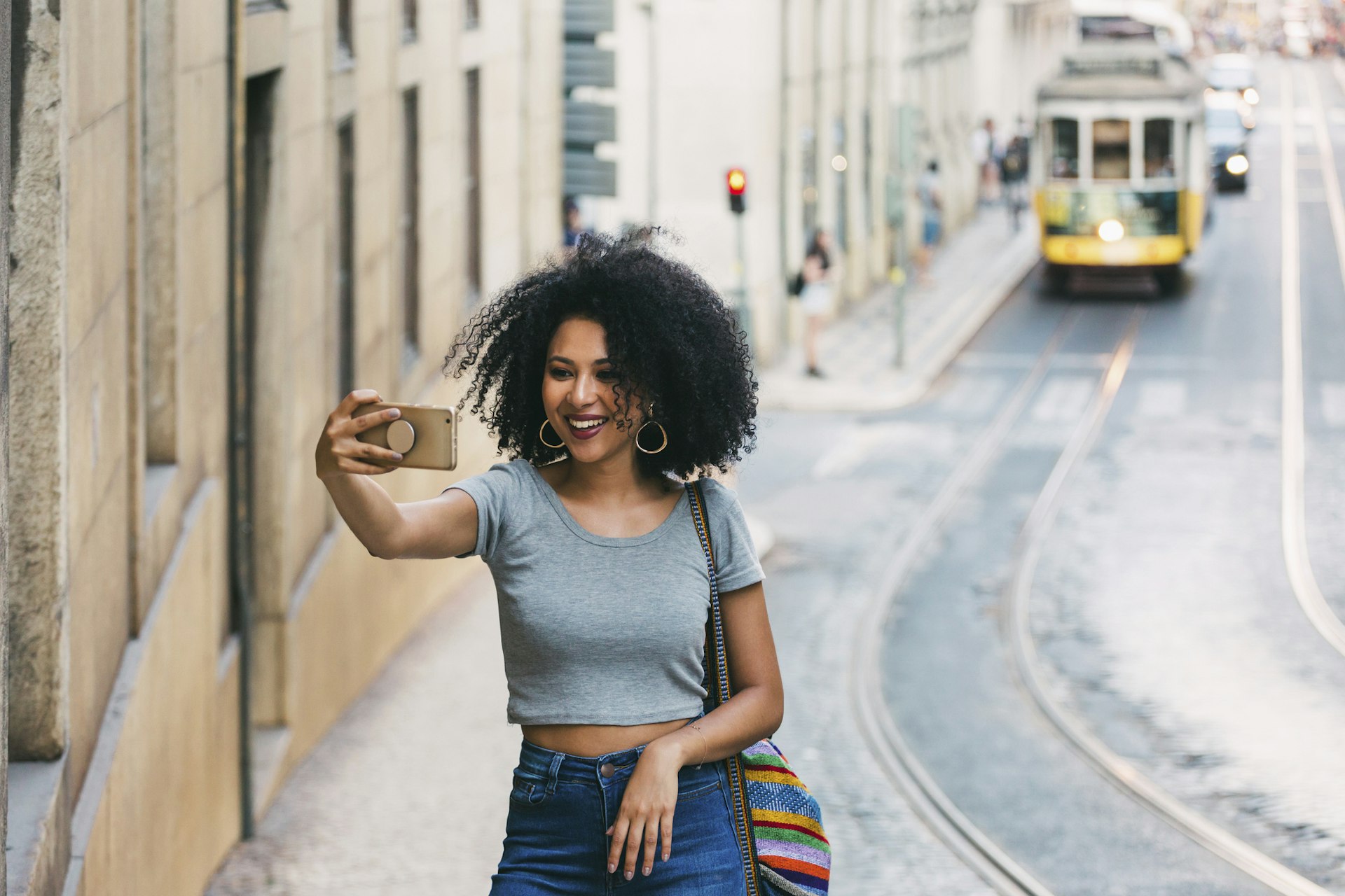 a woman smiles as she takes a selfie in Lisbon, Portugal as a yellow tram comes down the hill in the background