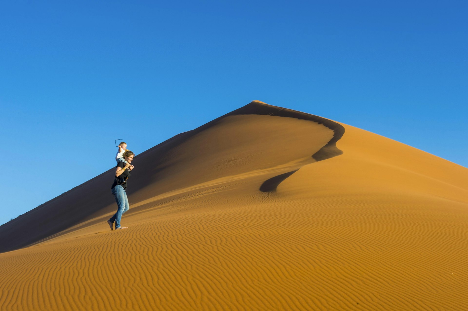 A woman carrying a baby on her shoulders walks along a steep ochre-colored sand dune that contrasts with the bright blue sky