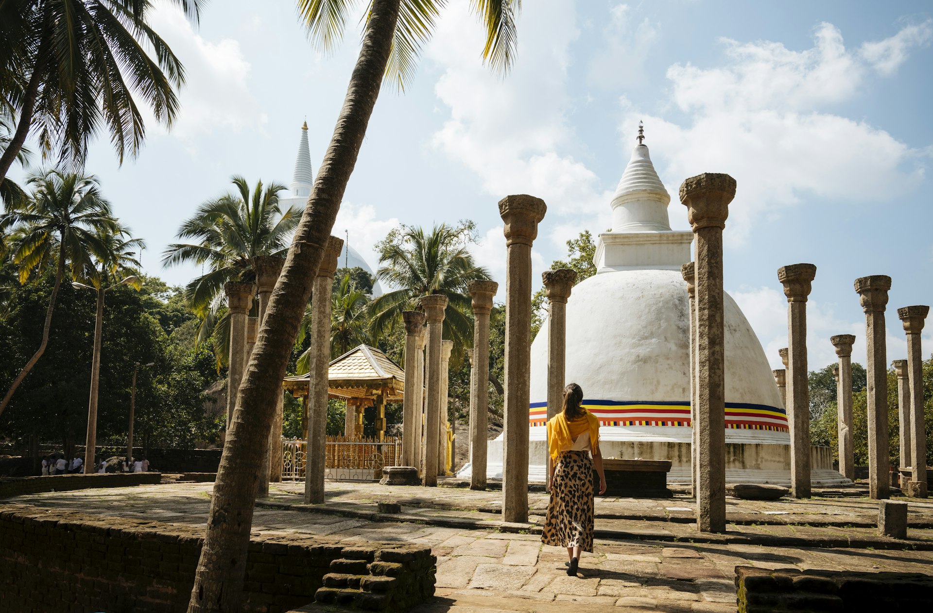 A woman shot from behind on a stone path, approaching a tipped-dome shrine (dagaba) ringed by columns in Sri Lanka 