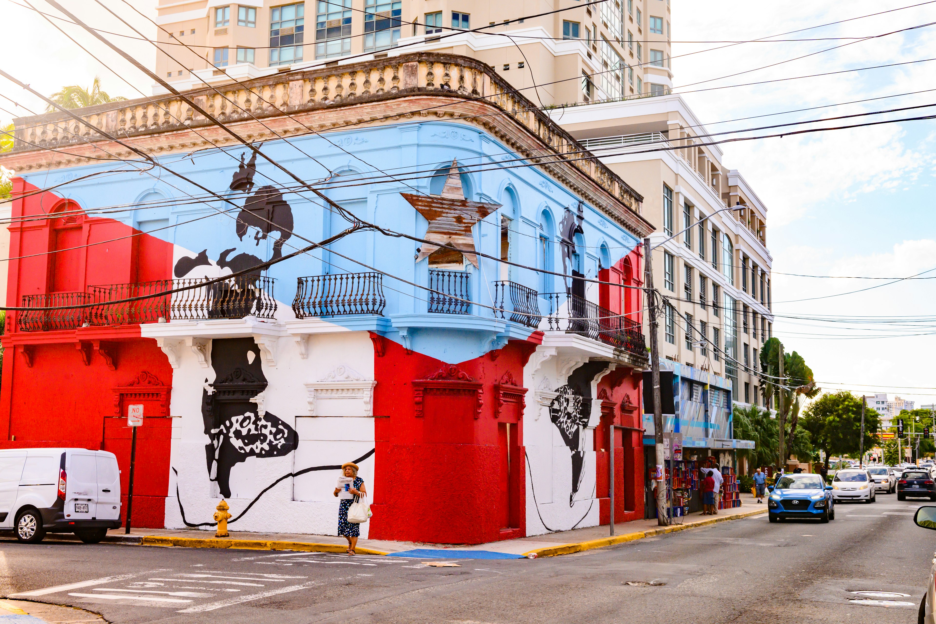 A street in Calle Loiza in the San Turce neighborhood of San Juan, Puerto Rico. A corner building is painted with the Puerto Rican flag. Cars and people can be seen along the street and sidewalk