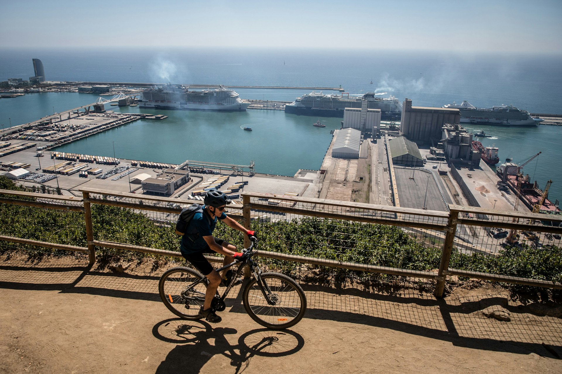 A cyclist rides along a viewpoint overlooking cruise ships docked at the Barcelona Cruise Terminal, Barcelona, Catalonia Spain