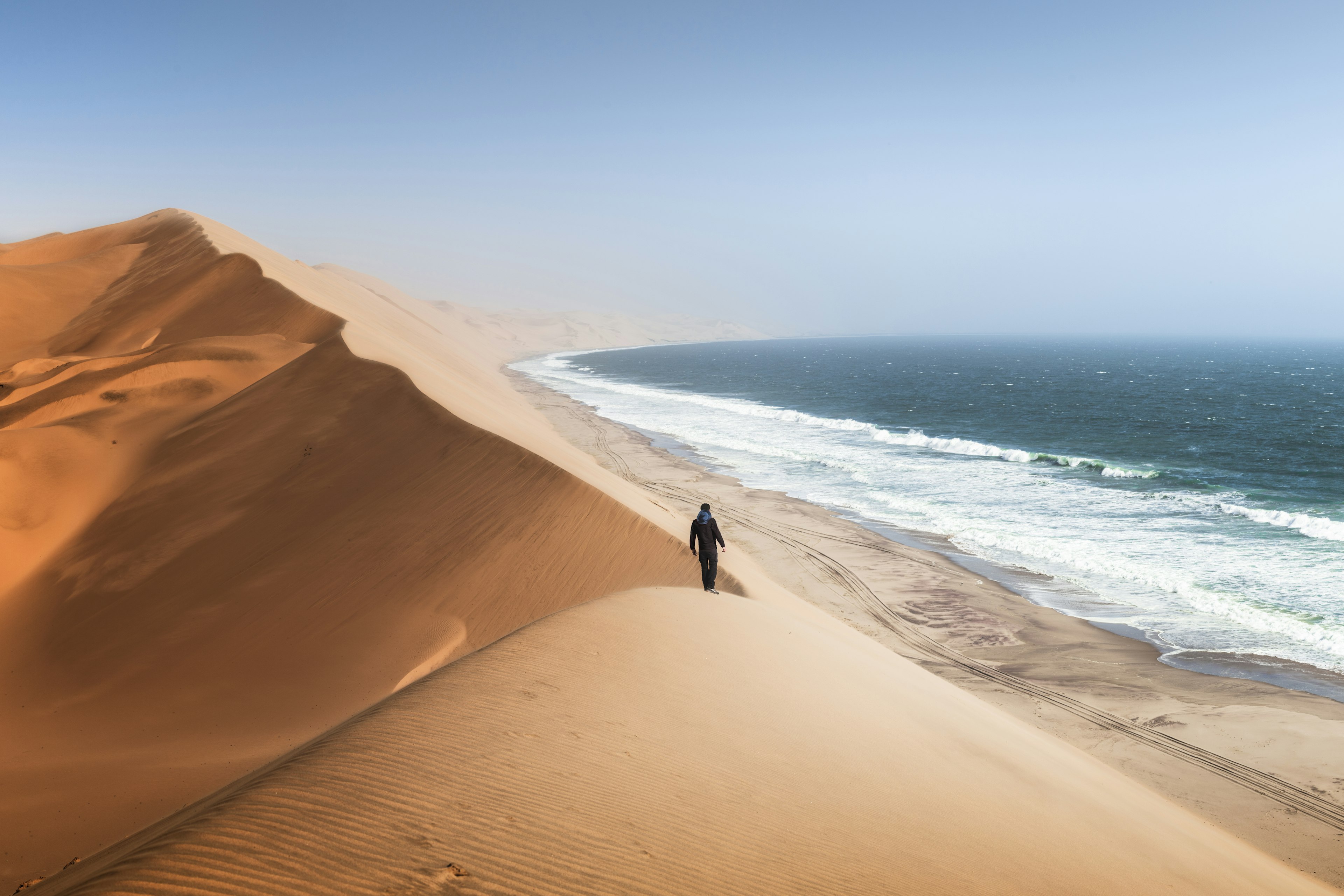 A person walks along the ridge of a large dune overlooking coastline to the right in Swakopmund, Namibia