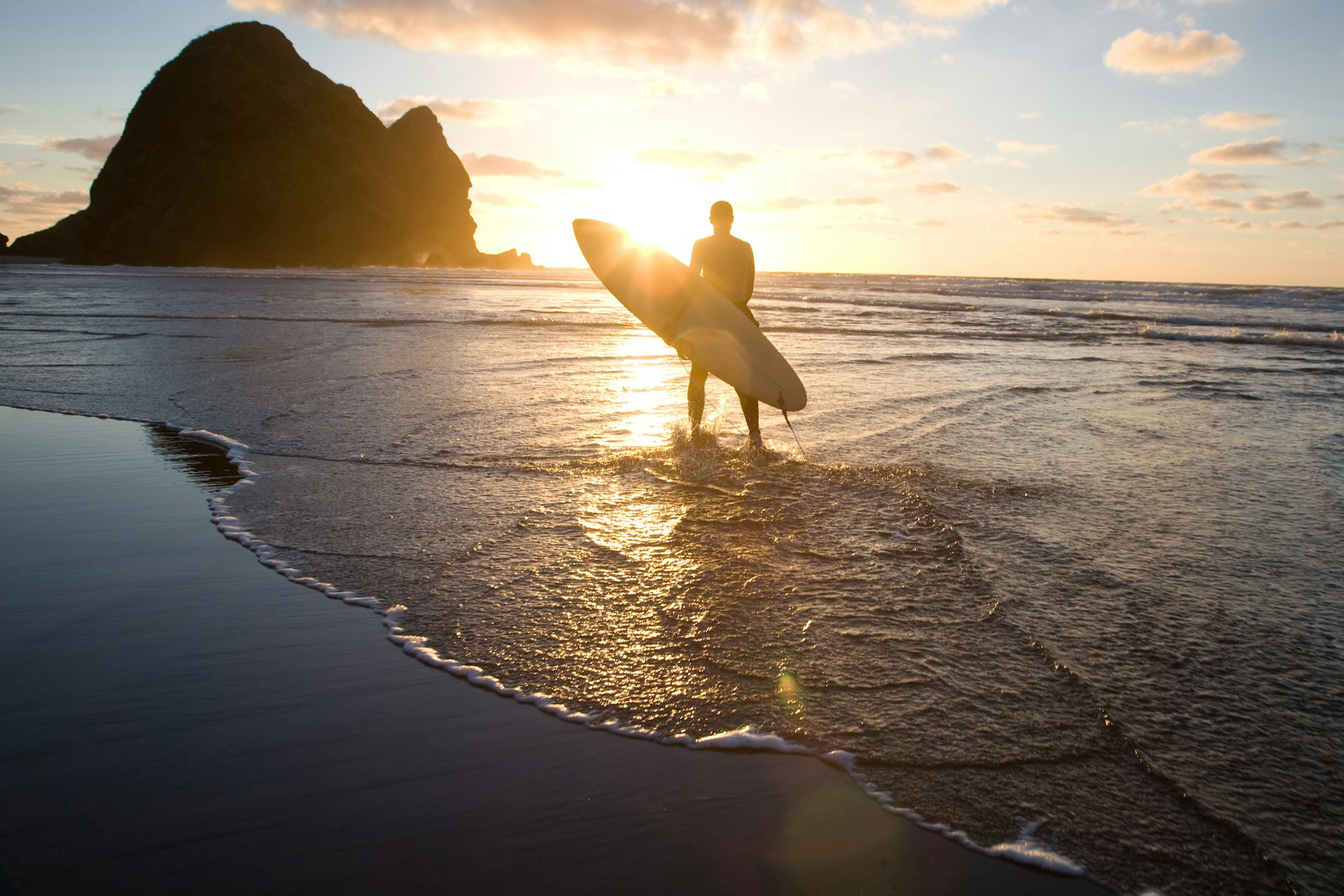 Silhouette of surfer holding surfboard and walking into water at sunset to surf, Piha Beach, Auckland, New Zealand.