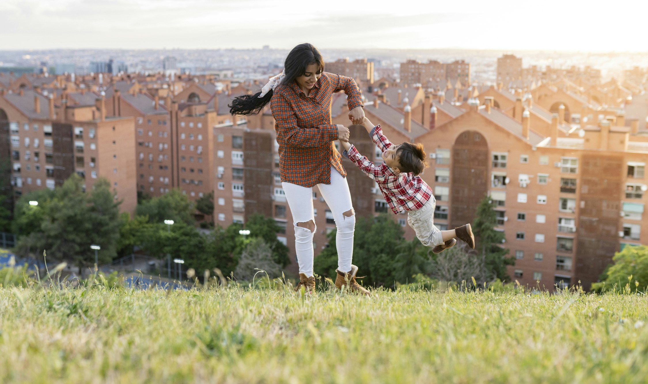 A mother swings her small boy around in an open space on a hill with a city in the background