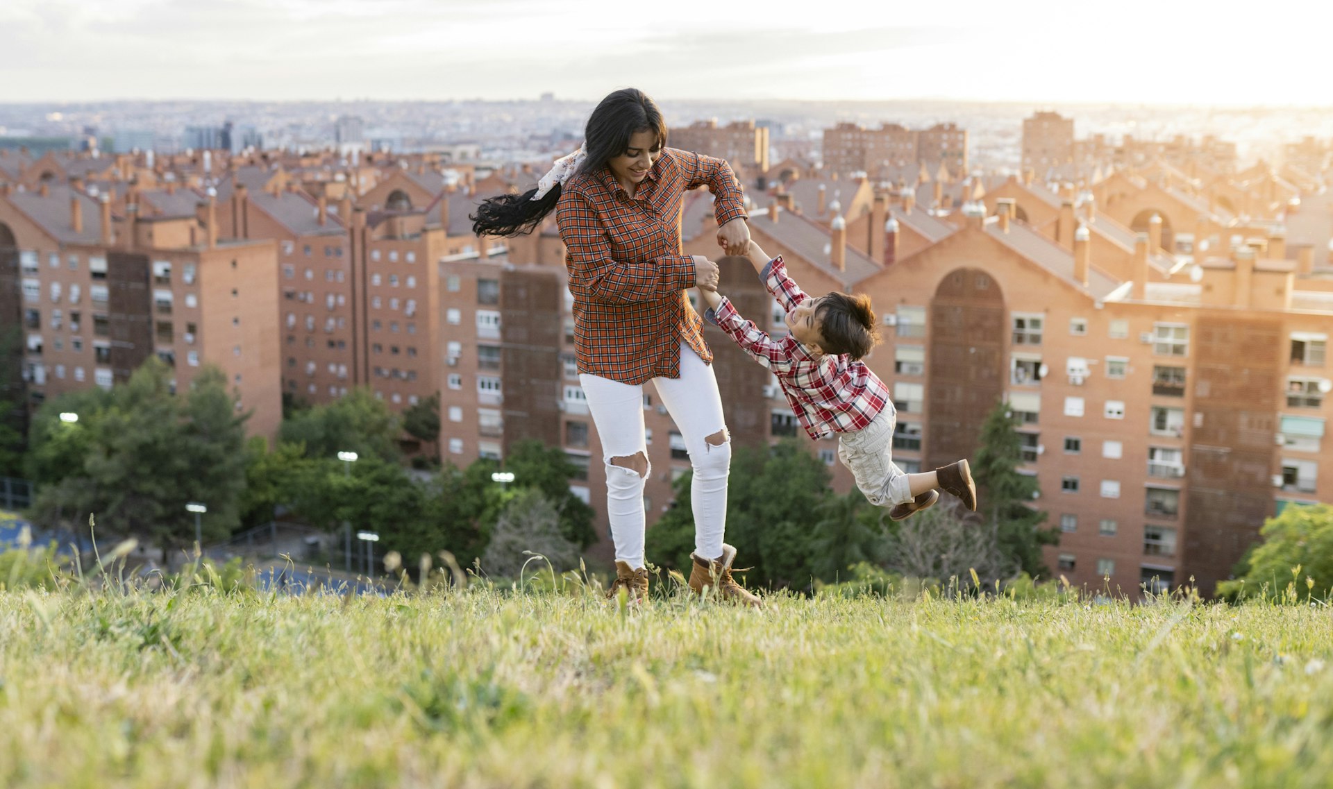 A mother swings her small boy around in an open space on a hill with a city in the background 