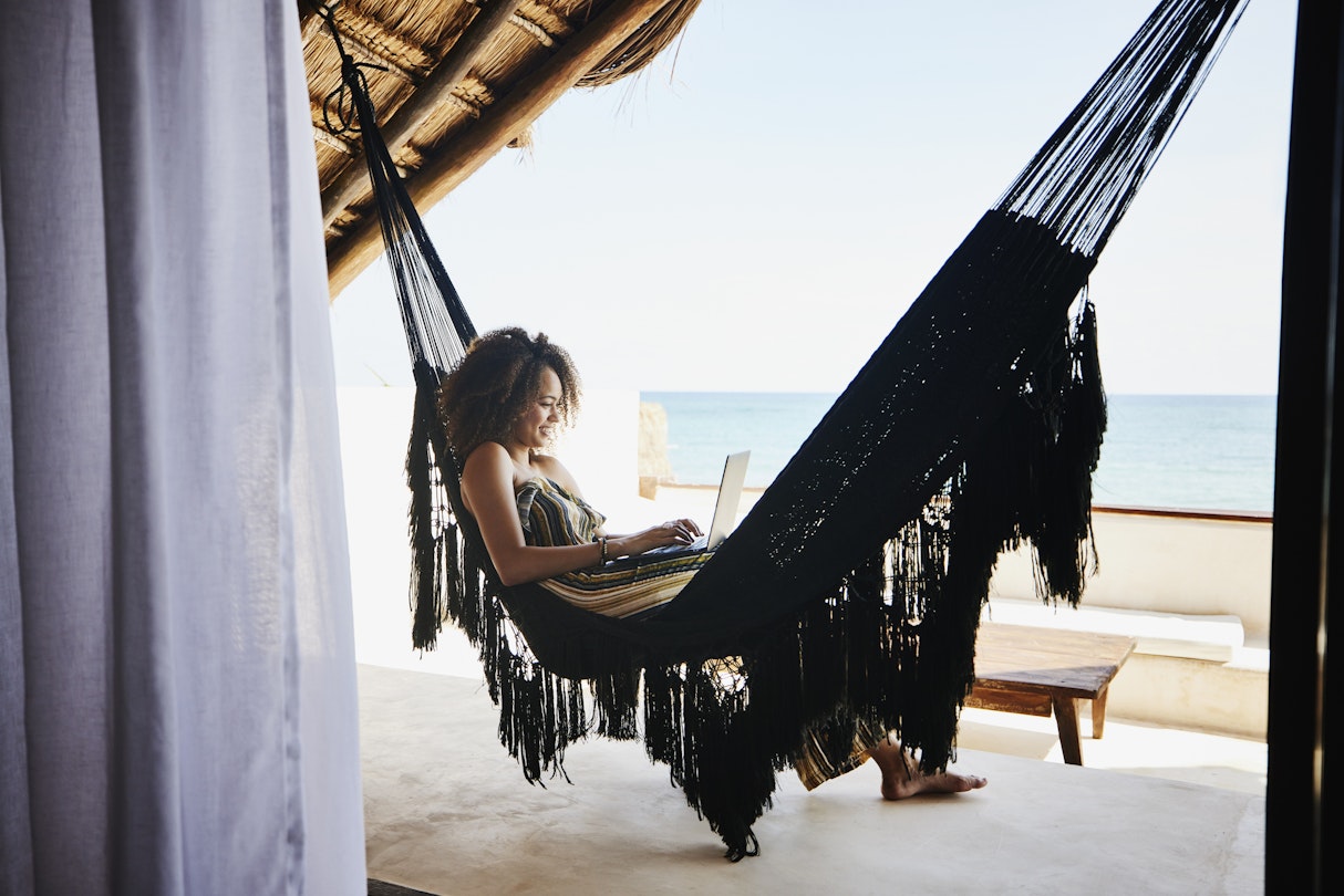 1366306539
Wide shot of a woman working on laptop while relaxing in a hammock in an ocean-view villa in Mexico