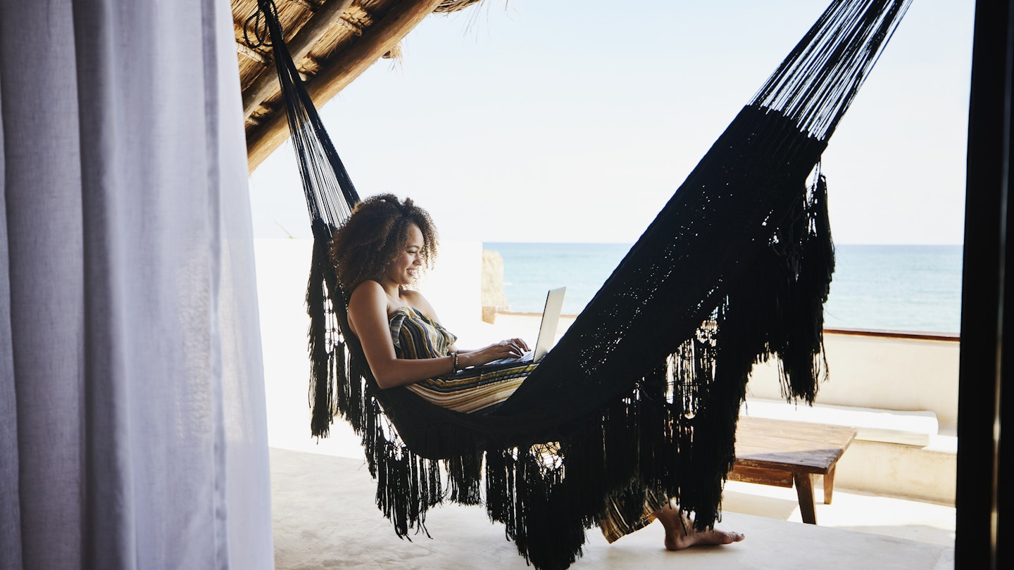 1366306539
Wide shot of a woman working on laptop while relaxing in a hammock in an ocean-view villa in Mexico