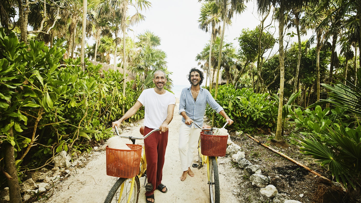 Two men laughing as they walk along a beach path with bikes in Mexico