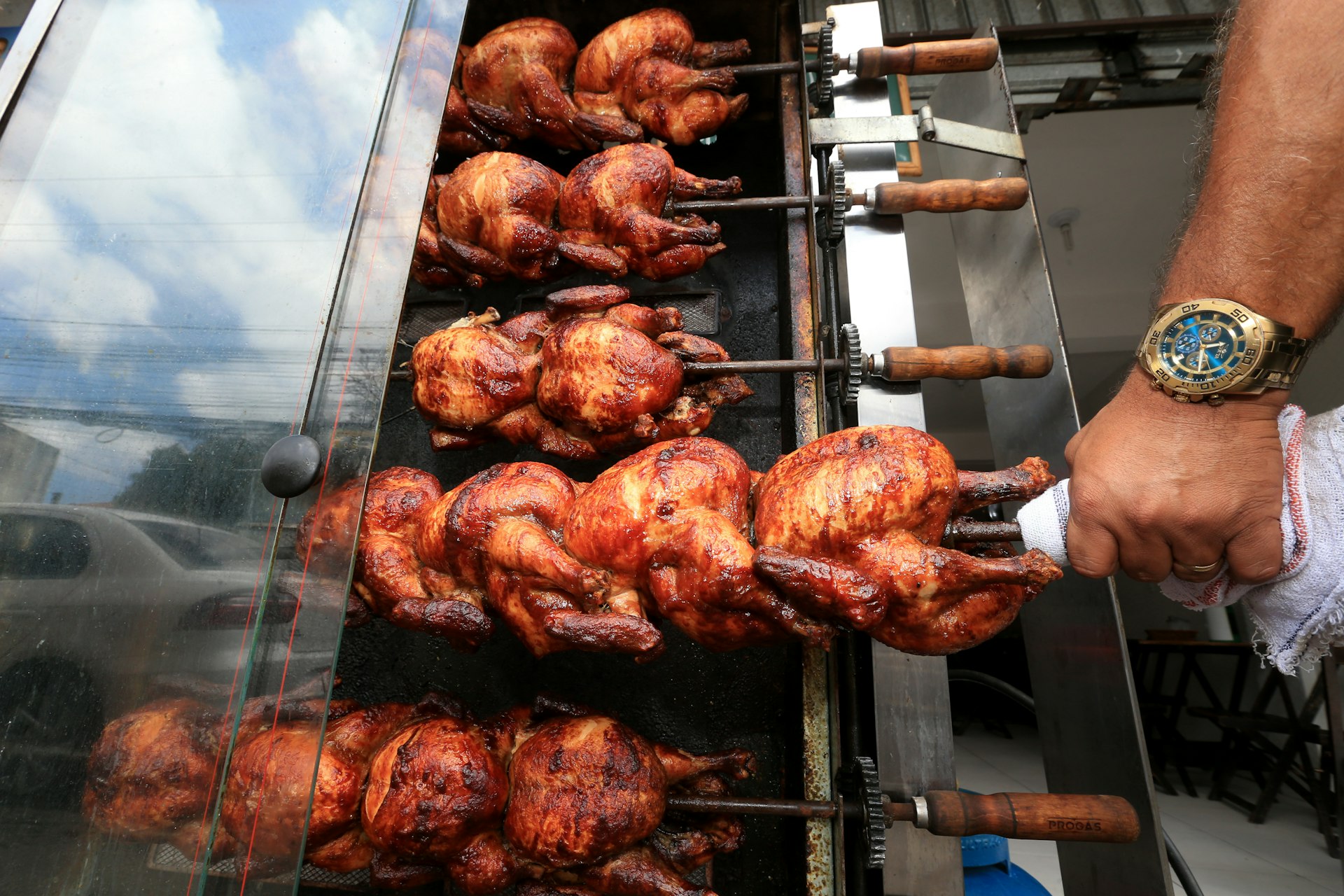 Rows of whole chickens on skewers roasting on a grill outside, a hand gripping a towel turning one of the skewers. At a street restaurant in Salvador, Brazil