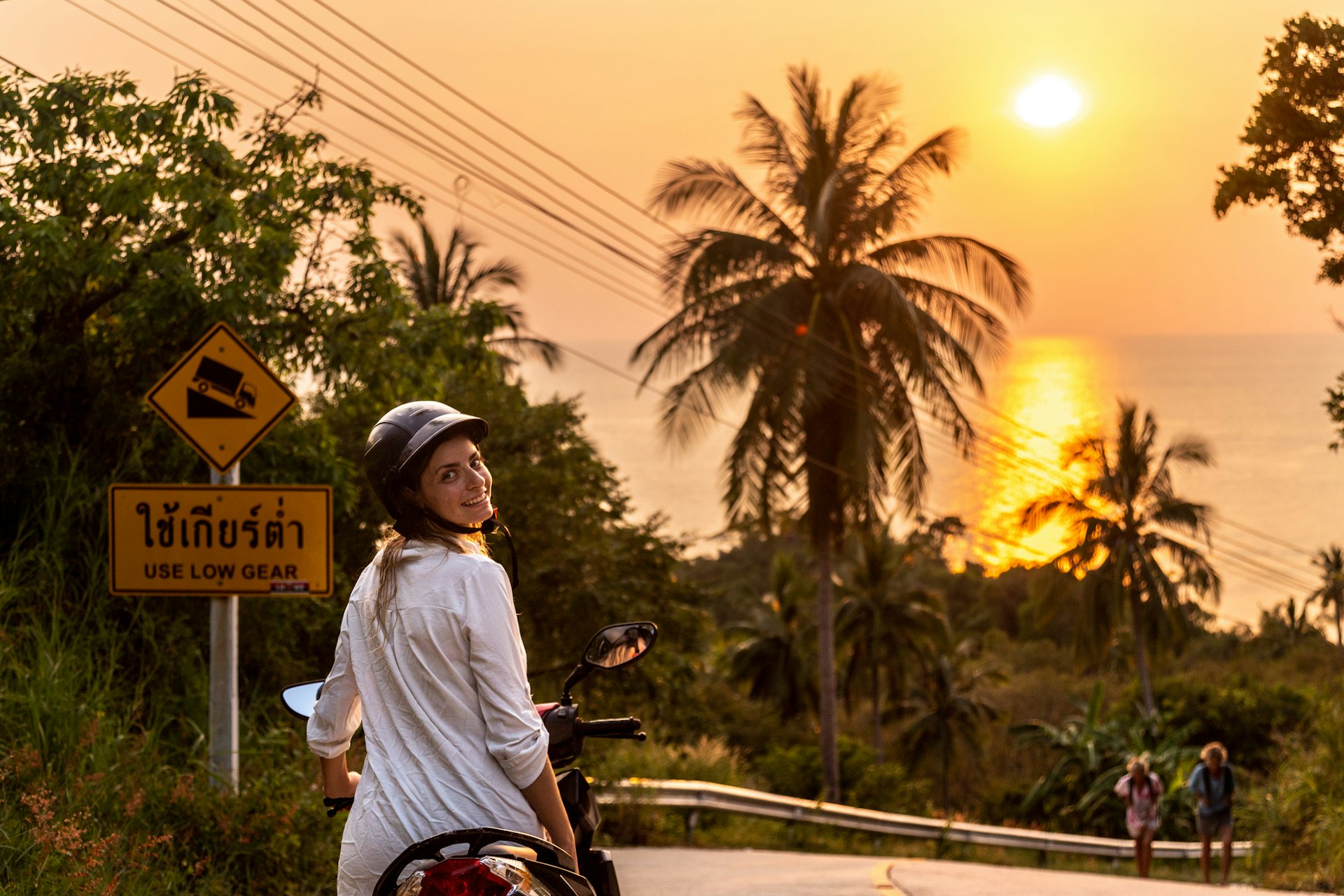 A woman in a helmet and sunglasses smiles at the camera as she rides a motorbike under palm trees in Thailand