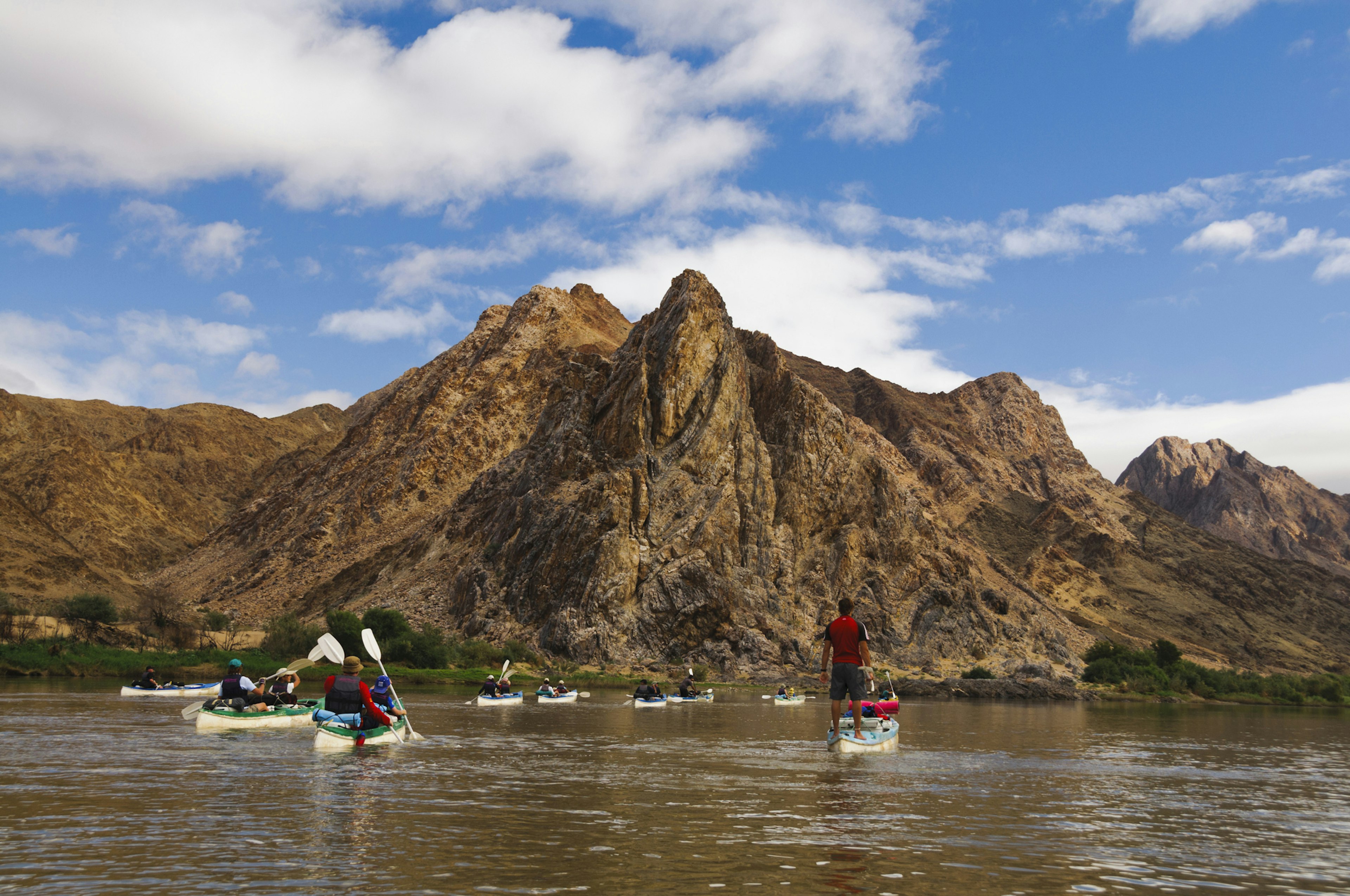 People paddle along in kayaks on a river with large rocky peaks in the surrounding landscape