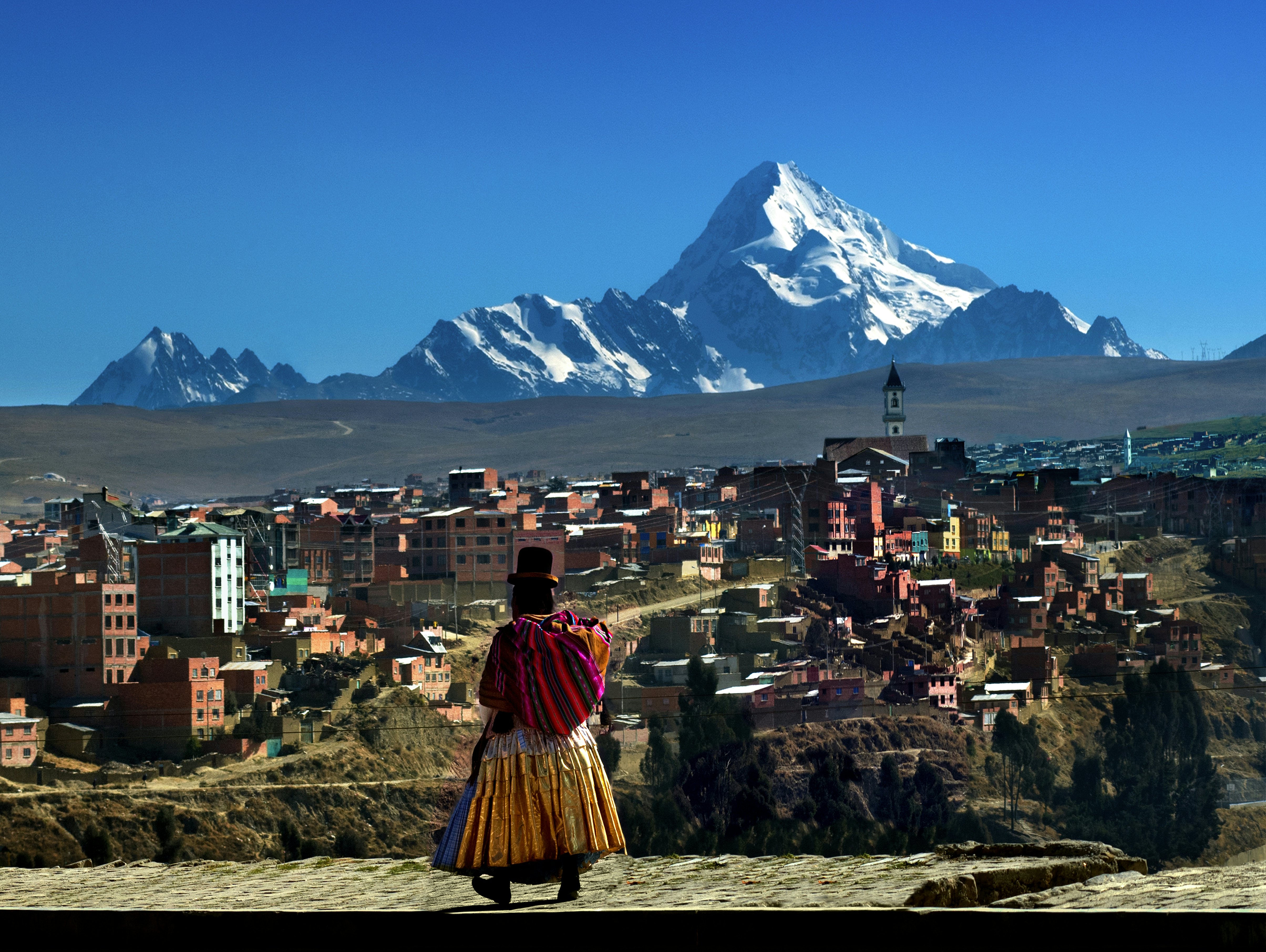 A woman in traditional Bolivian dress walks towards a small city with a snowcapped mountain looming over it