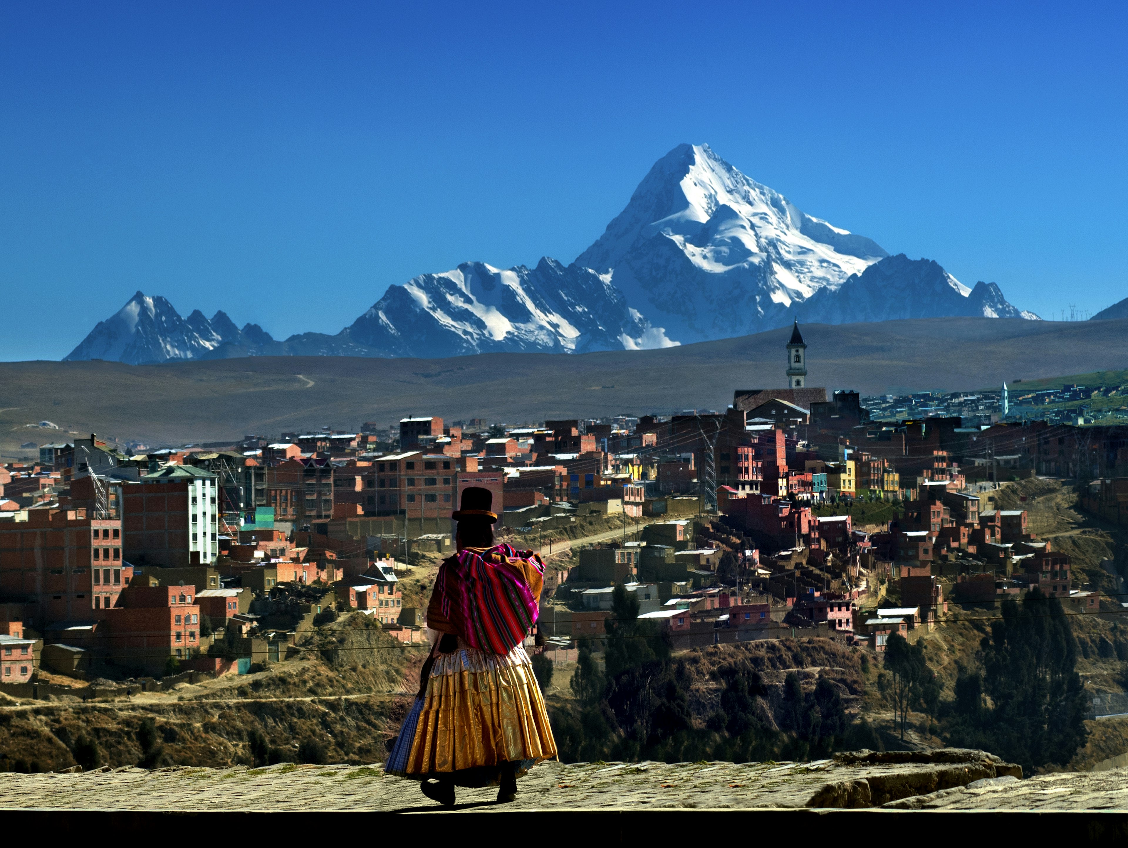 A woman in traditional Bolivian dress walks towards a small city with a snowcapped mountain looming over it