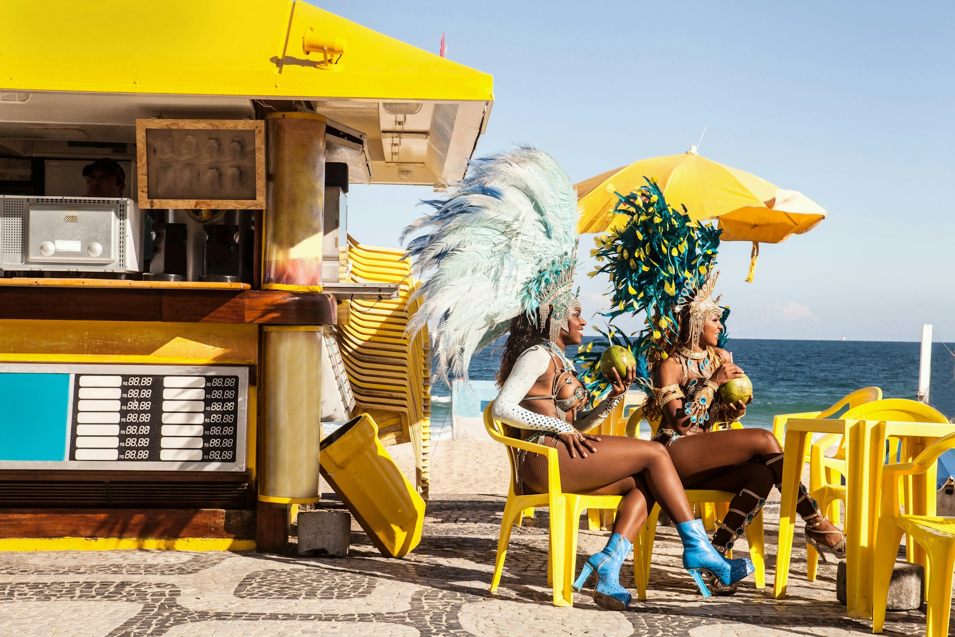 This image features two samba dancers taking a break at Ipanema Beach in Rio de Janeiro. They are seated on yellow chairs under a yellow umbrella, adjacent to a snack bar with a blue and yellow facade. The dancers are adorned with elaborate carnival costumes with feathers in hues of blue, green, and yellow. They appear relaxed, enjoying coconuts, with the ocean in the background and a clear blue sky above. The scene captures the vibrant and festive atmosphere typical of Rio's Carnaval.