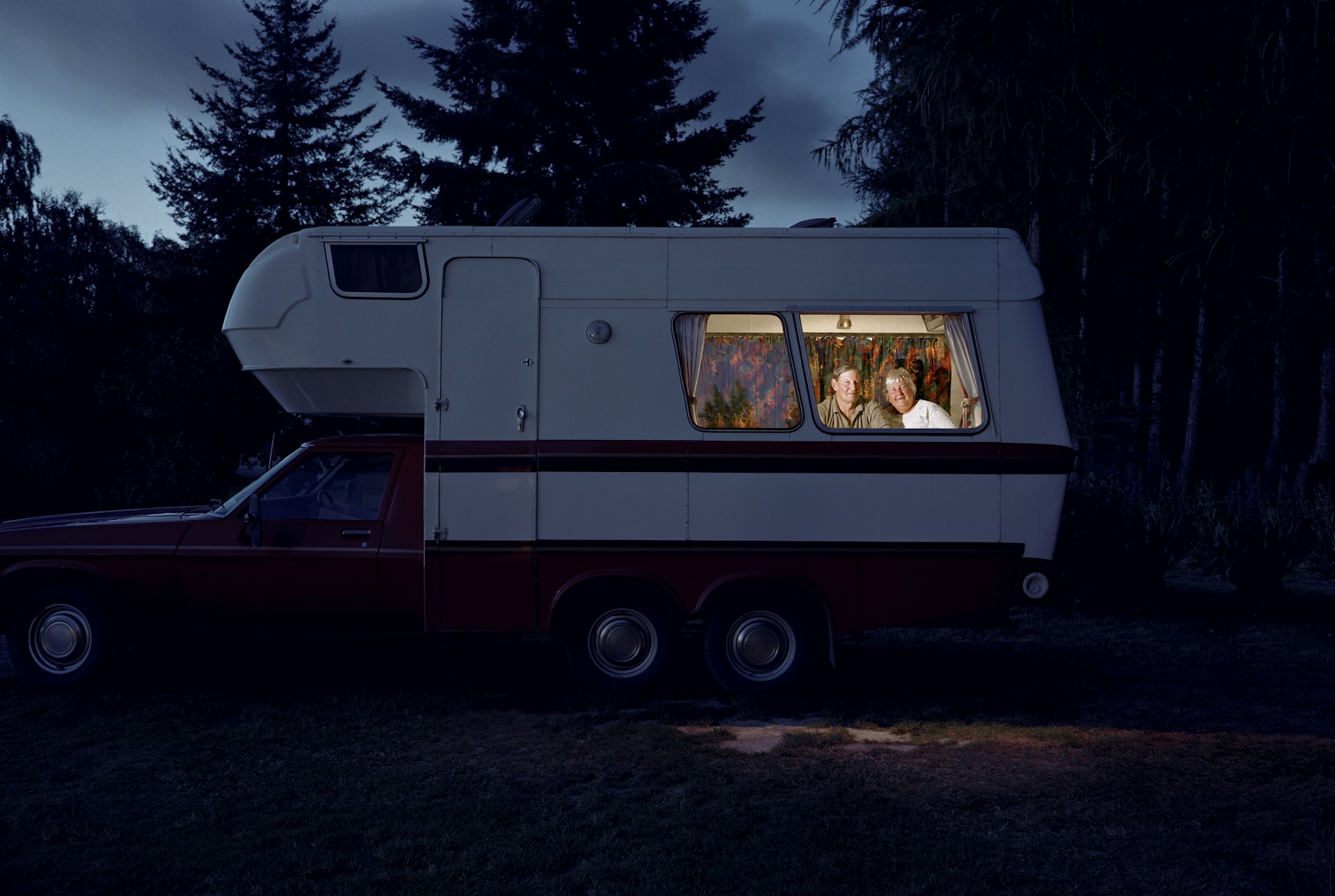 An elderly couple sitting inside their brightly-lit campervan are seen through the window with the Auckland darkness outside