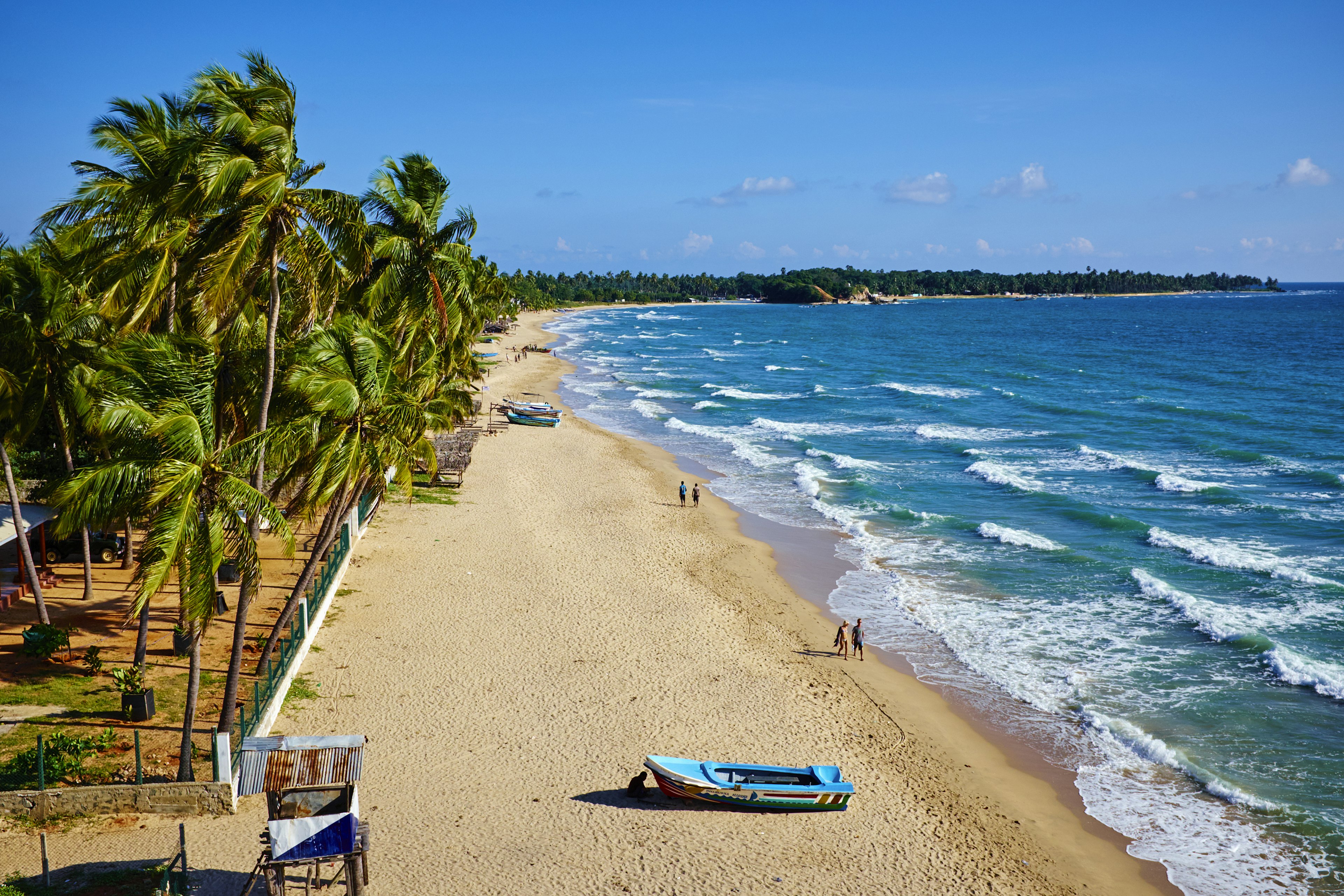 A scenic aerial shot of a mostly empty beach with palm trees, golden sand and gentle turquoise waves, two couples walking by the water and a small boat on the sand in the foreground