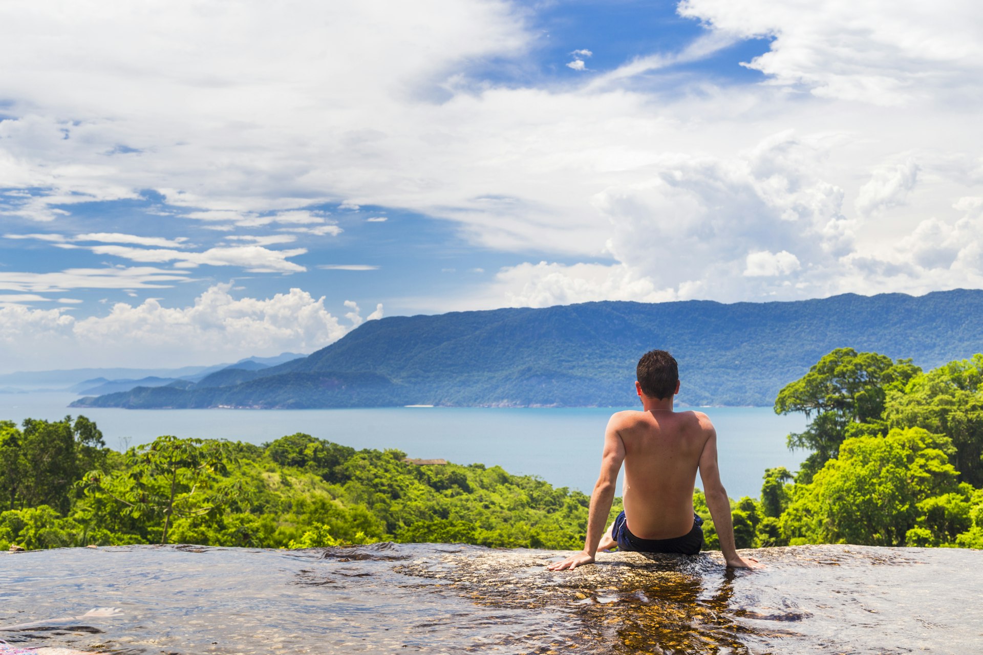 Young shirtless man, looking at view of green hills, blue water, and grey cloud-covered hills in the distance