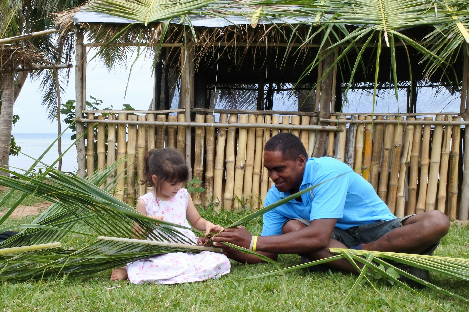 A Fijian man shows a young tourist girl how to create a basket from weaving together palm fronds