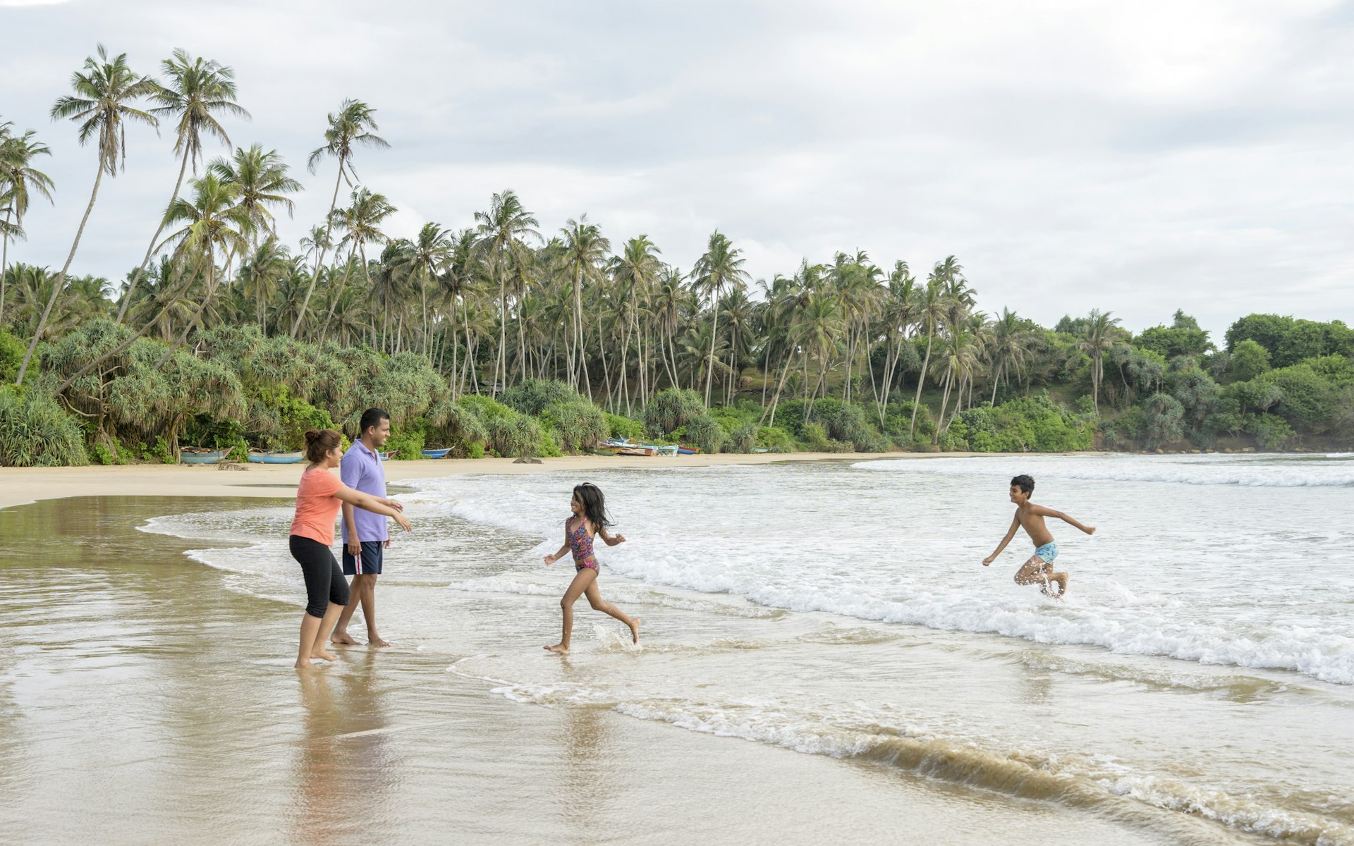 Two young children run out of the sea towards their parents on a sandy beach backed by palm trees