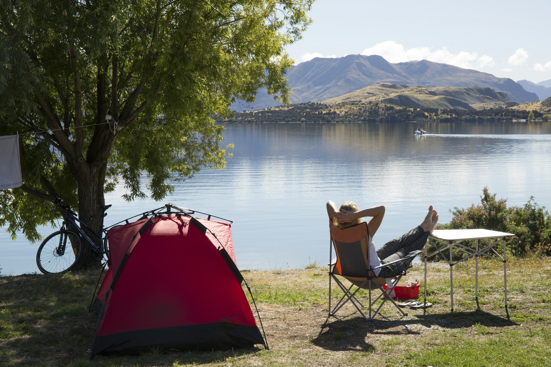 Woman at a lakeside campsite seen in silhouette from behind, sitting back with her legs up and crossed in front of her and her arms on her head, beside a tent and a bike 