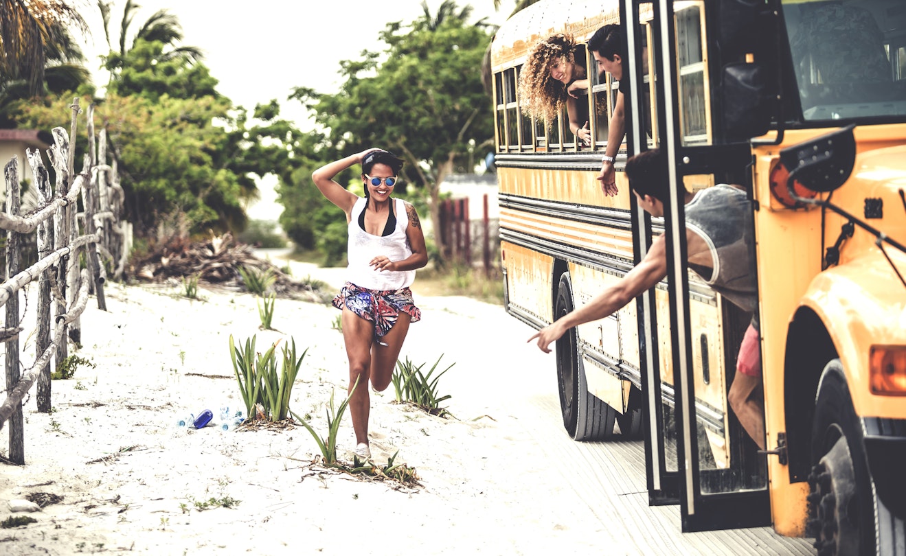 A young woman running up to an old yellow school bus as her friends wave