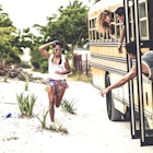 A young woman running up to an old yellow school bus as her friends wave