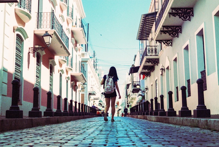 Girl Walking Down Cobblestone Street Surrounded By Colorful Buildings