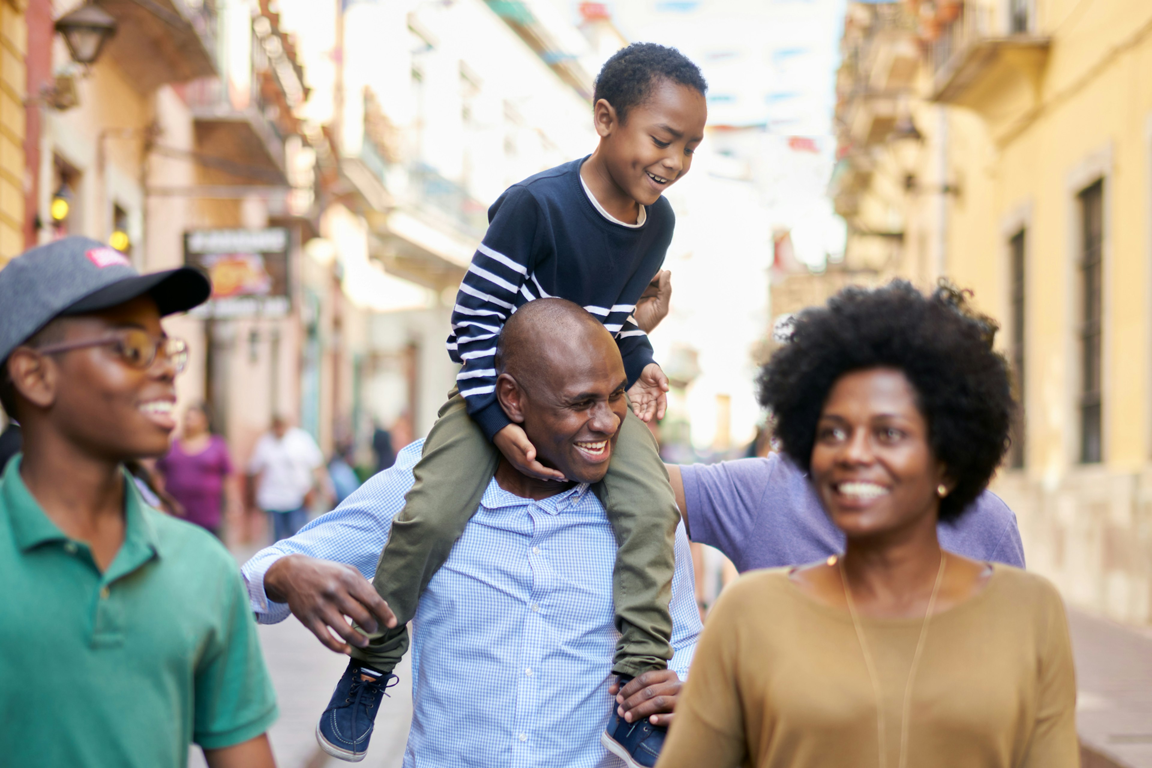 A Black family walks down the street in Mexico City, the father carrying the younger son on his shoulders and the mother and teenage son walking ahead and smiling.