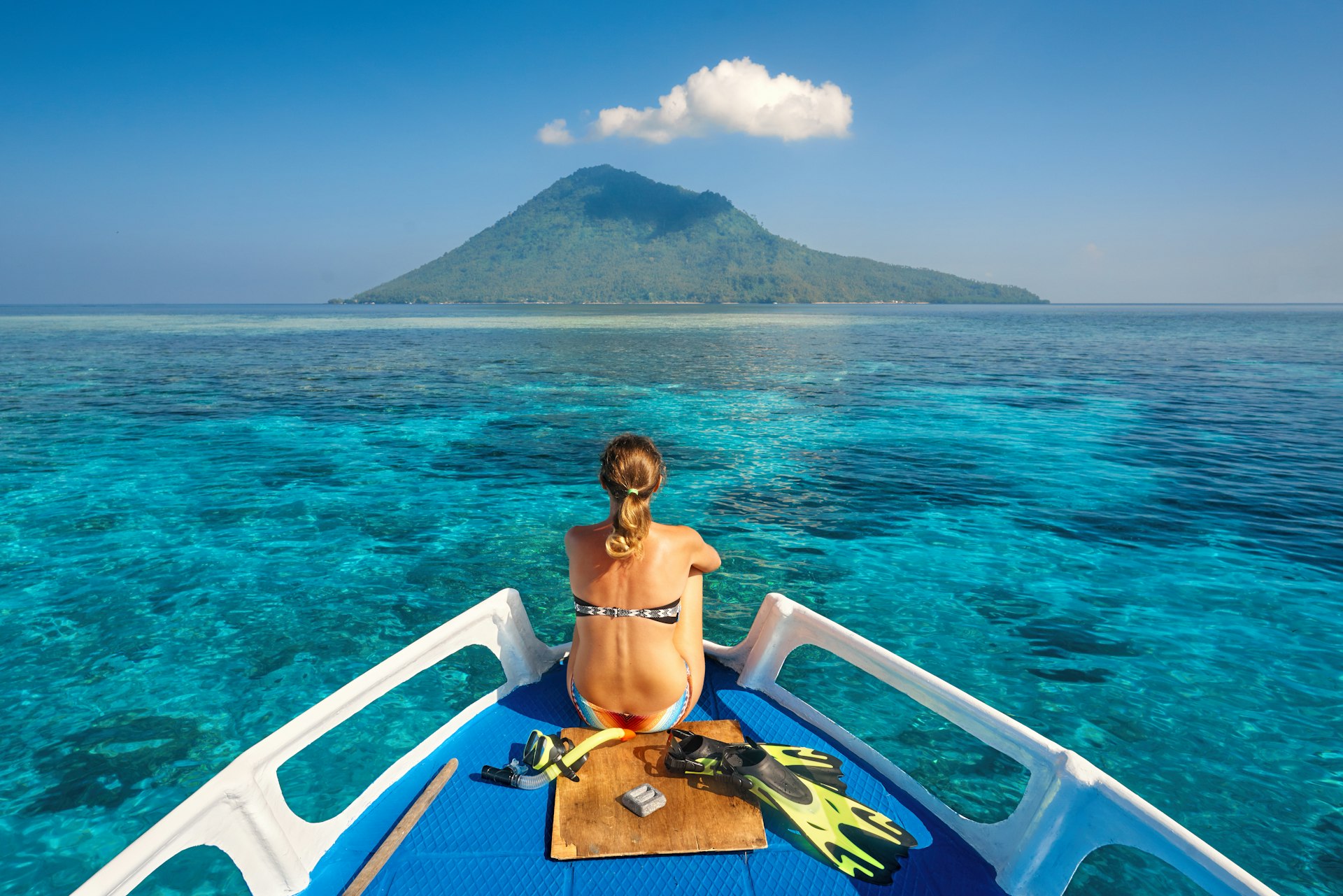 Young woman in swimsuit sat on a boat with a mask and flippers looking to a clean sea and volcano Manado Tua. North Sulawesi, Indonesia