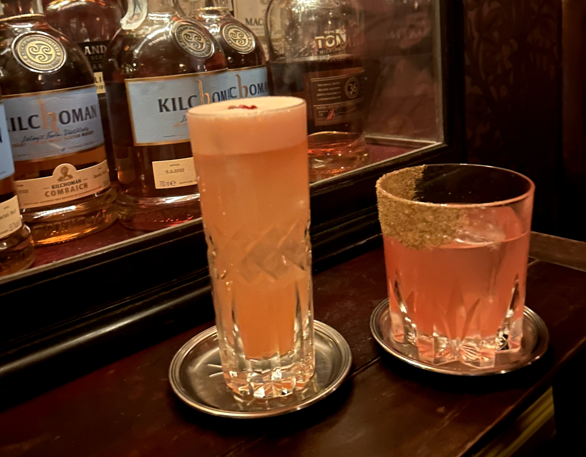 Two cocktail drinks on display in front of a drinks cabinet with several bottle of spirits in them
