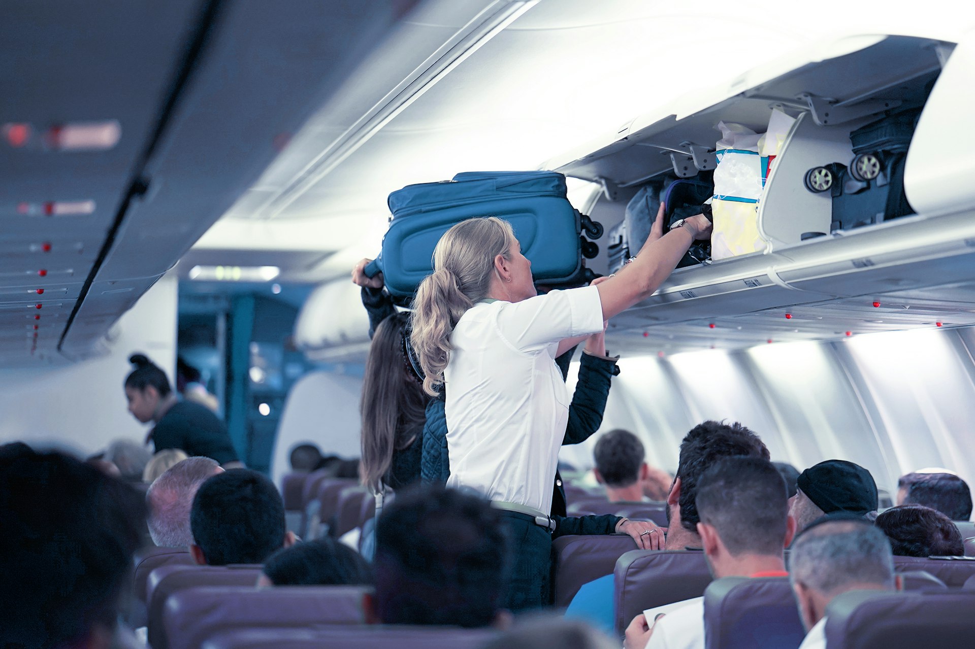 A flight attendant helps passengers load baggage in the overhead compartments of an airplane
