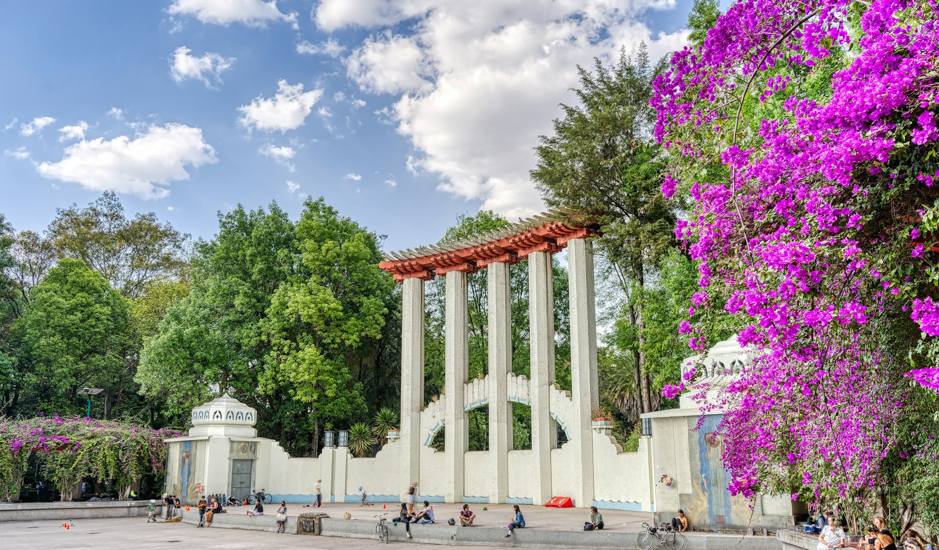 People sitting in a park in front of a giant row of columns and fucshia flowers