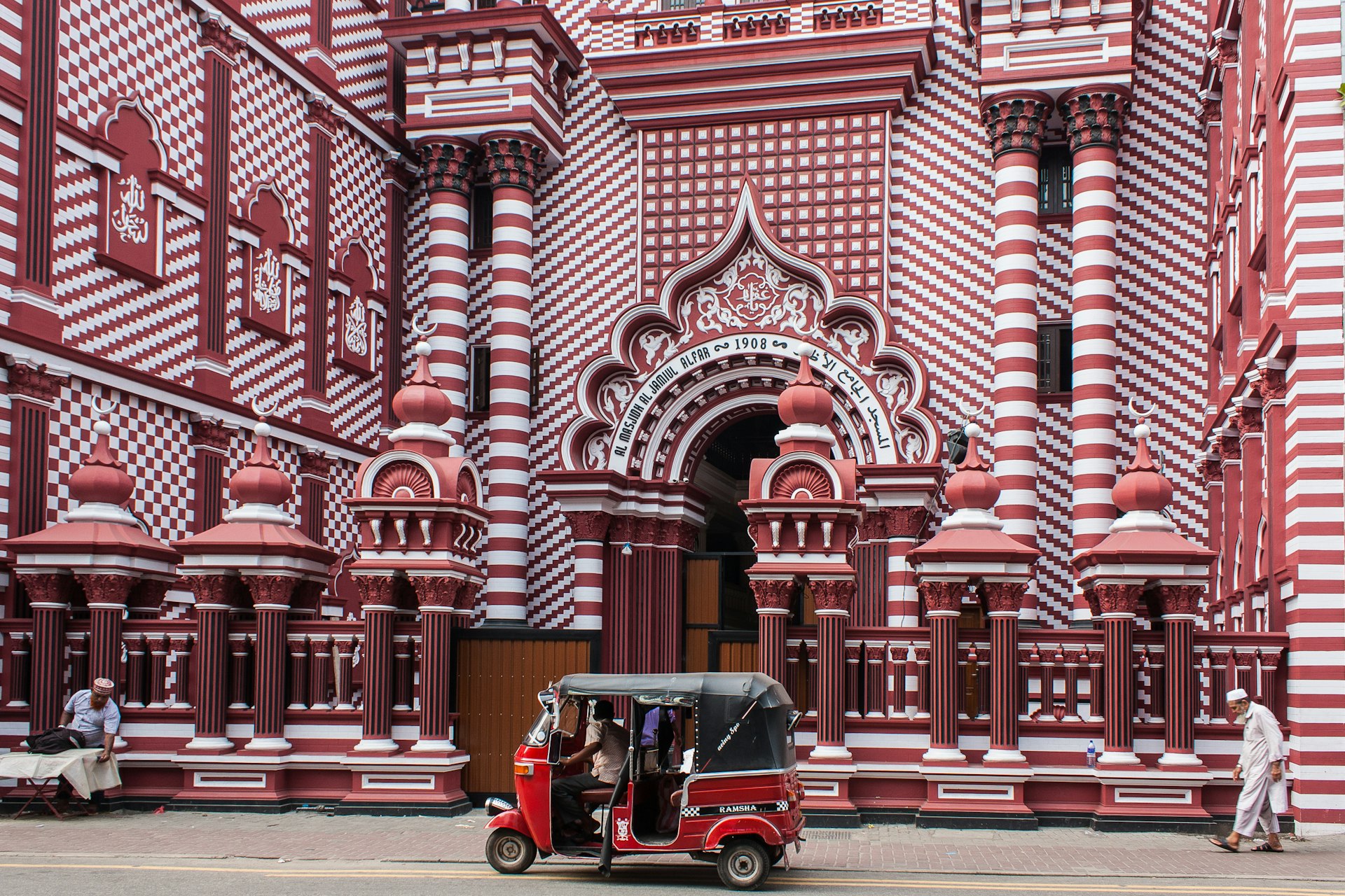 A red auto rickshaw, a pedestrian, and a vendor setting up in front of the decorative red-and-white patterned facade of Jamiul Alfar Mosque, built in 1908, in the heart of the bazaar of Pettah, one of the oldest districts in Colombo