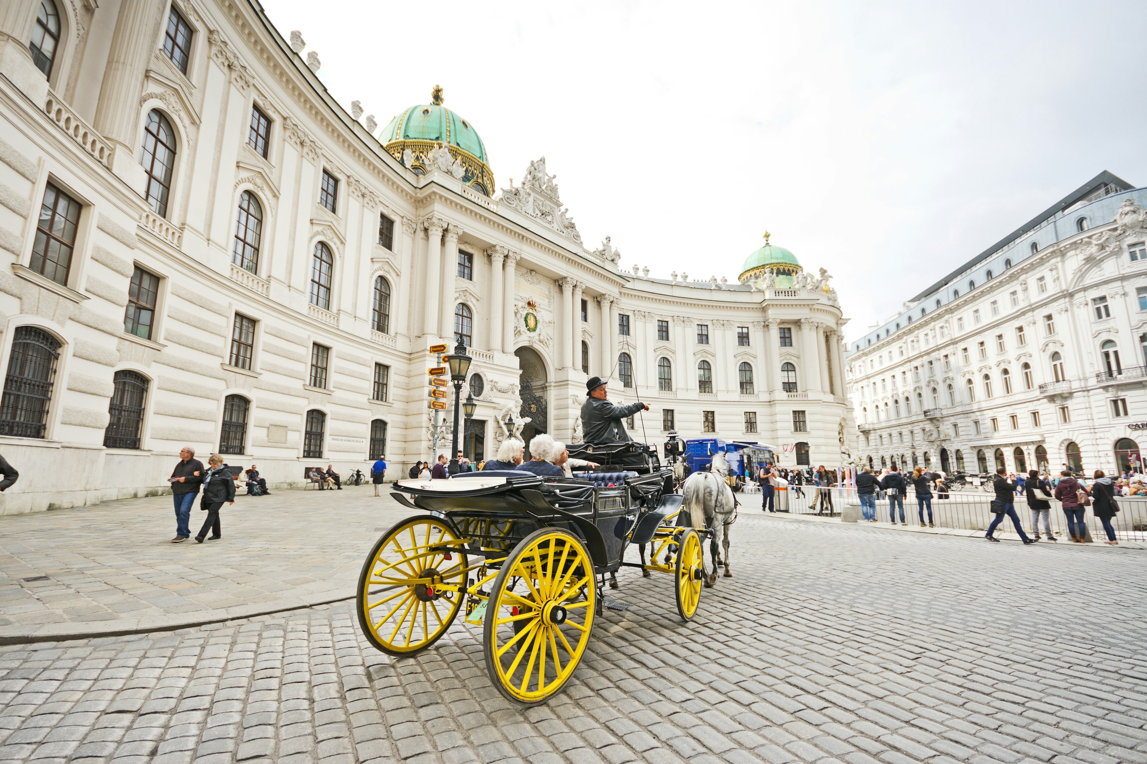 A horse-drawn carriage called fiaker with coachman as tourist guide passing by the Hofburg palace at Michaelerplatz with tourists and people