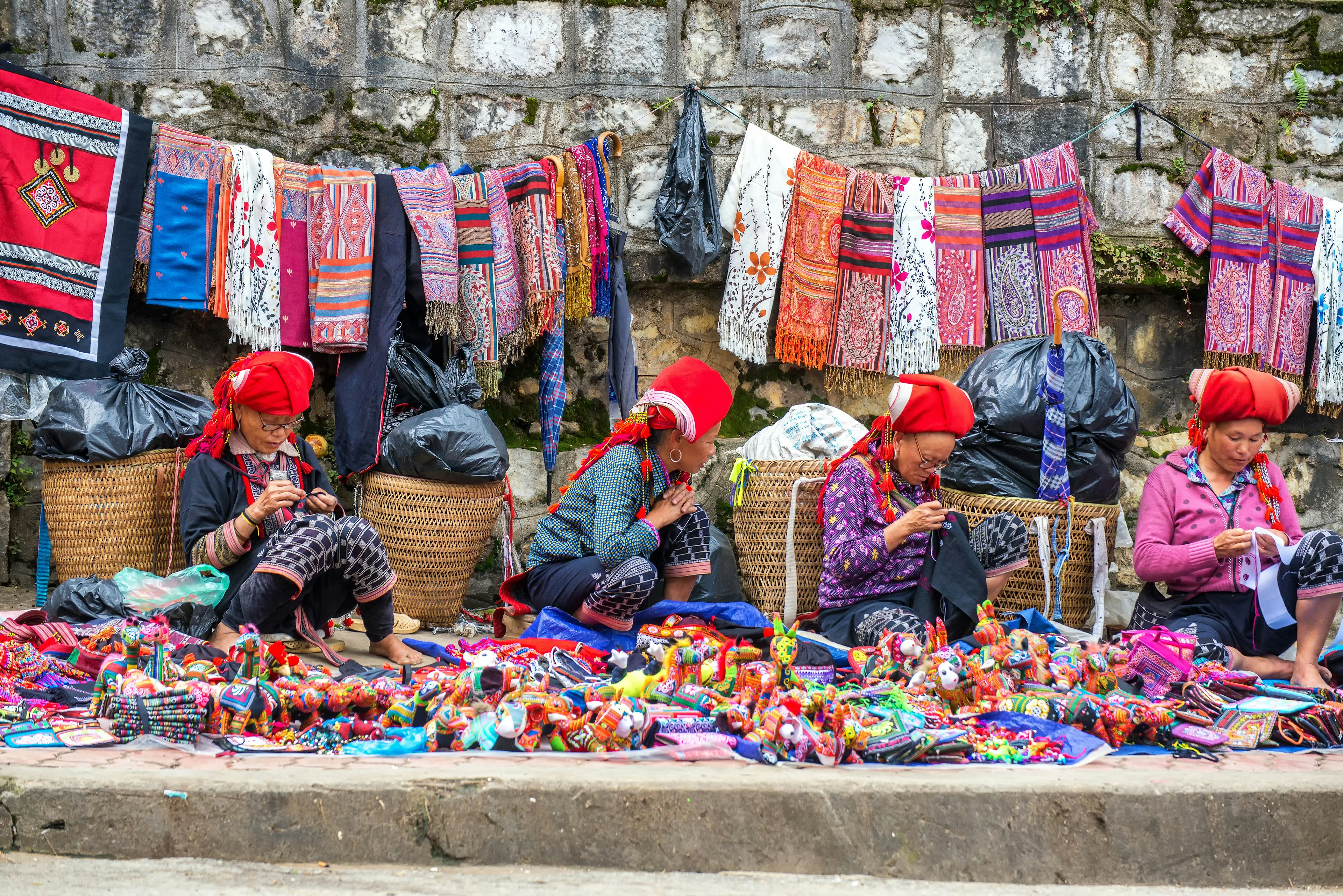 Four Hmong Women Sit in Front of Baskets as They Work on Handcrafts. Traditional Woven Fabrics Hang on the Rock Wall Behind Thema, and more Goods Are Spread in Front of THE Main Street in Sapa, Vietnam