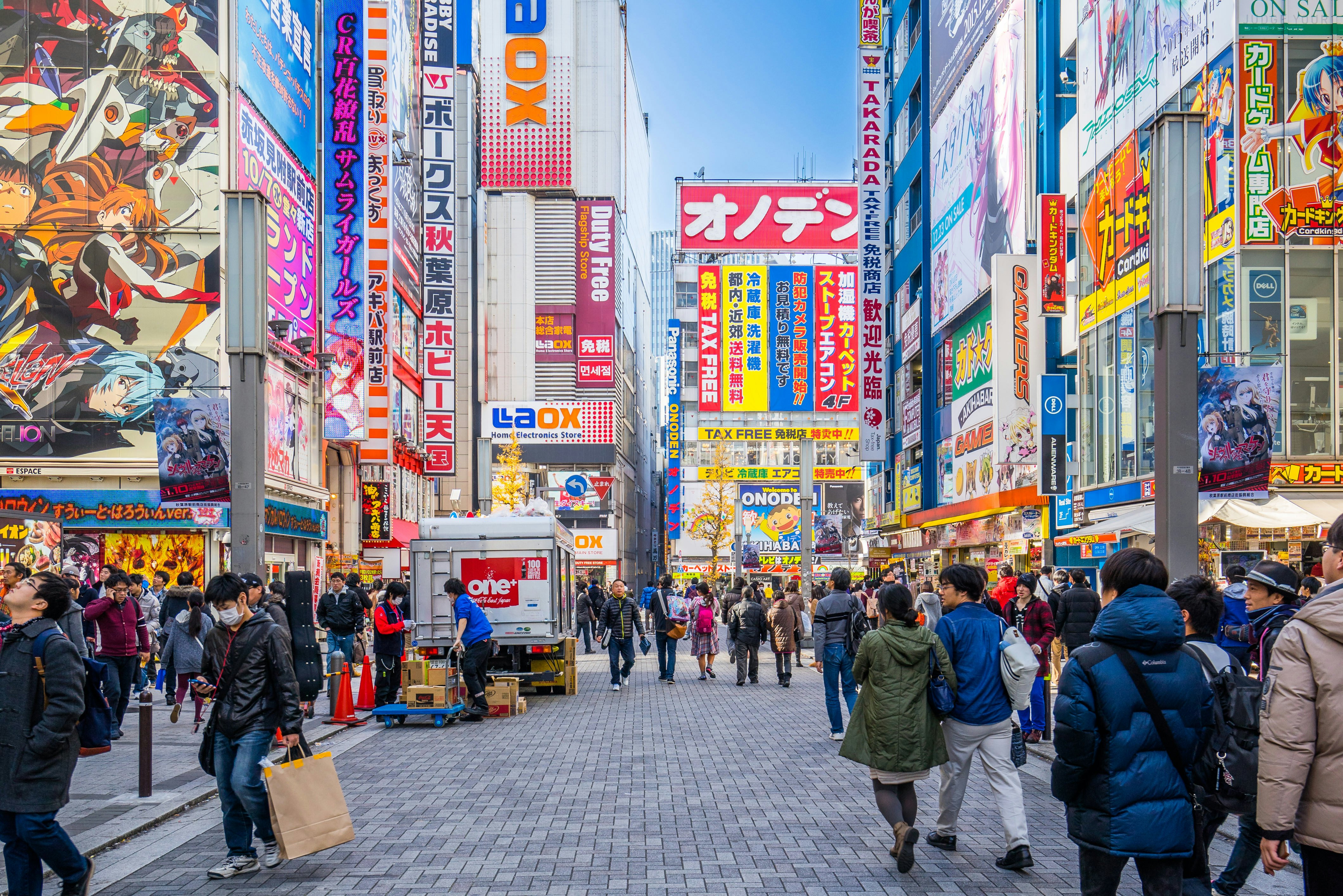 Shoppers walk along a narrow street in Akihabara, tall buildings covered with bright signs on either side