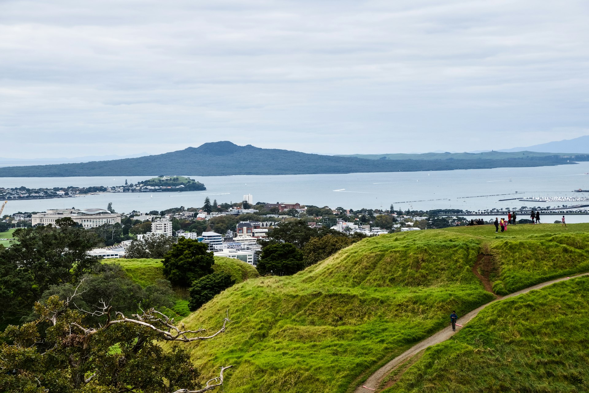 A grass-covered volcanic mound with several walkers on it on the edge of a bay 