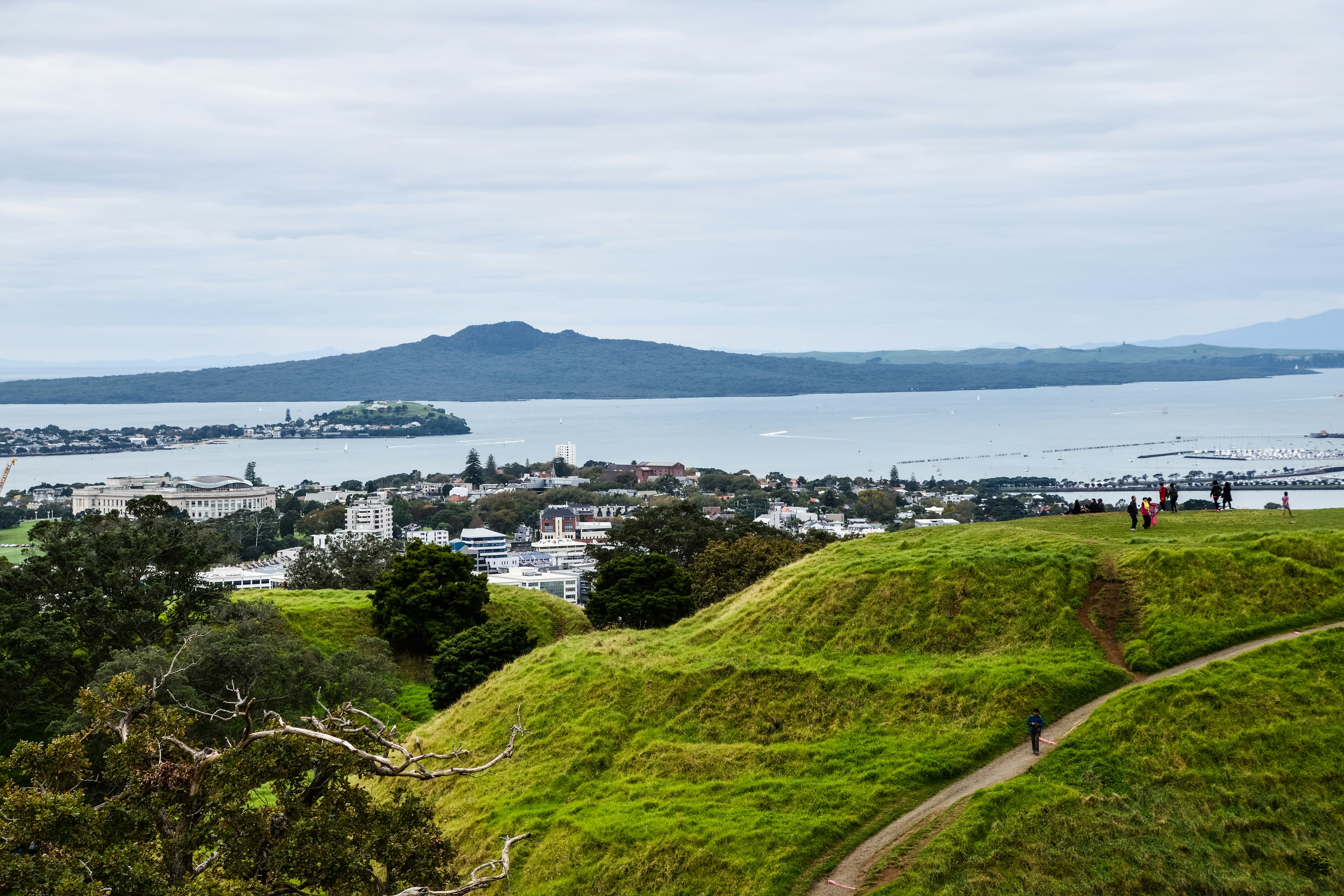 A grass-covered volcanic mound with several walkers on it on the edge of a bay