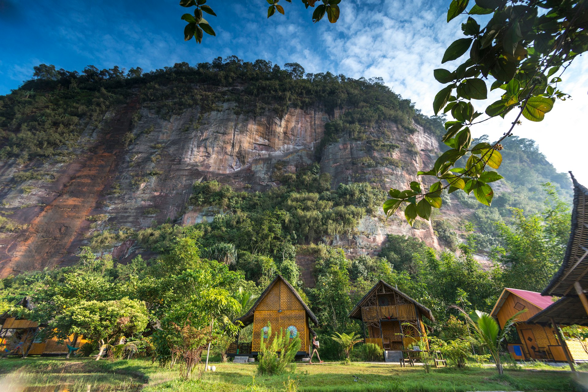 A series of wooden huts stand at the foot of a large cliff in a jungle